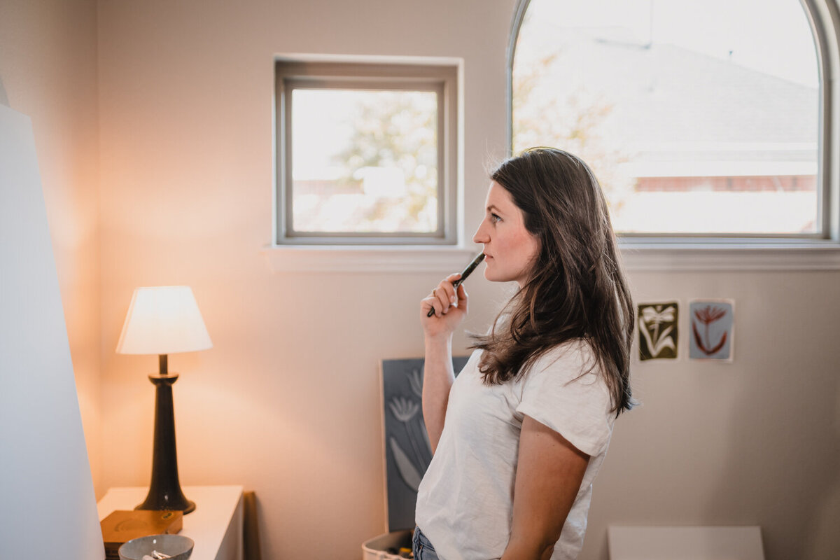 exas artist holding brush to lips while thoughtfully looking at canvas on easel during in-home branding session.