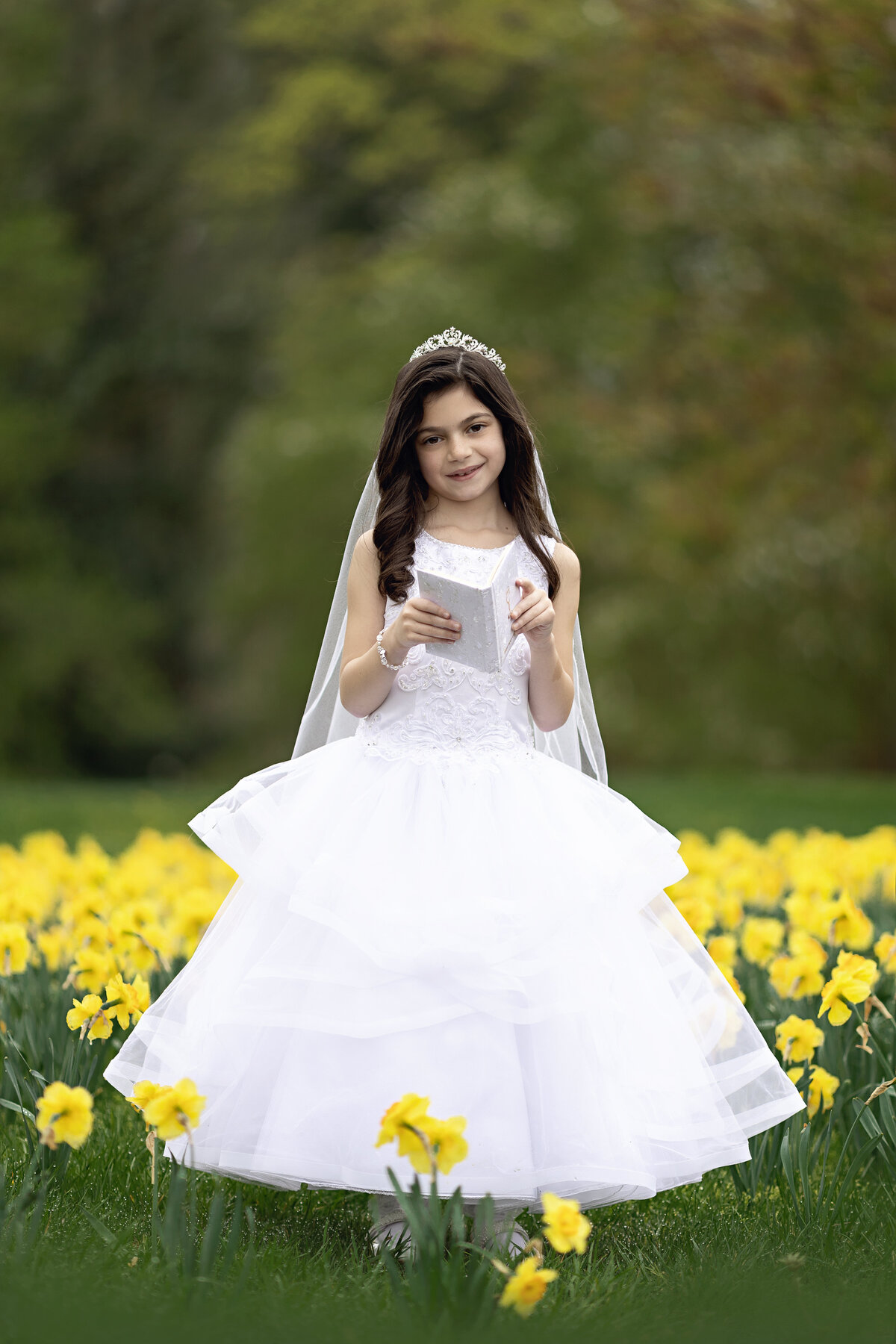 A girl with dark hair reads a book while standing among yellow daffodils in a long white dress