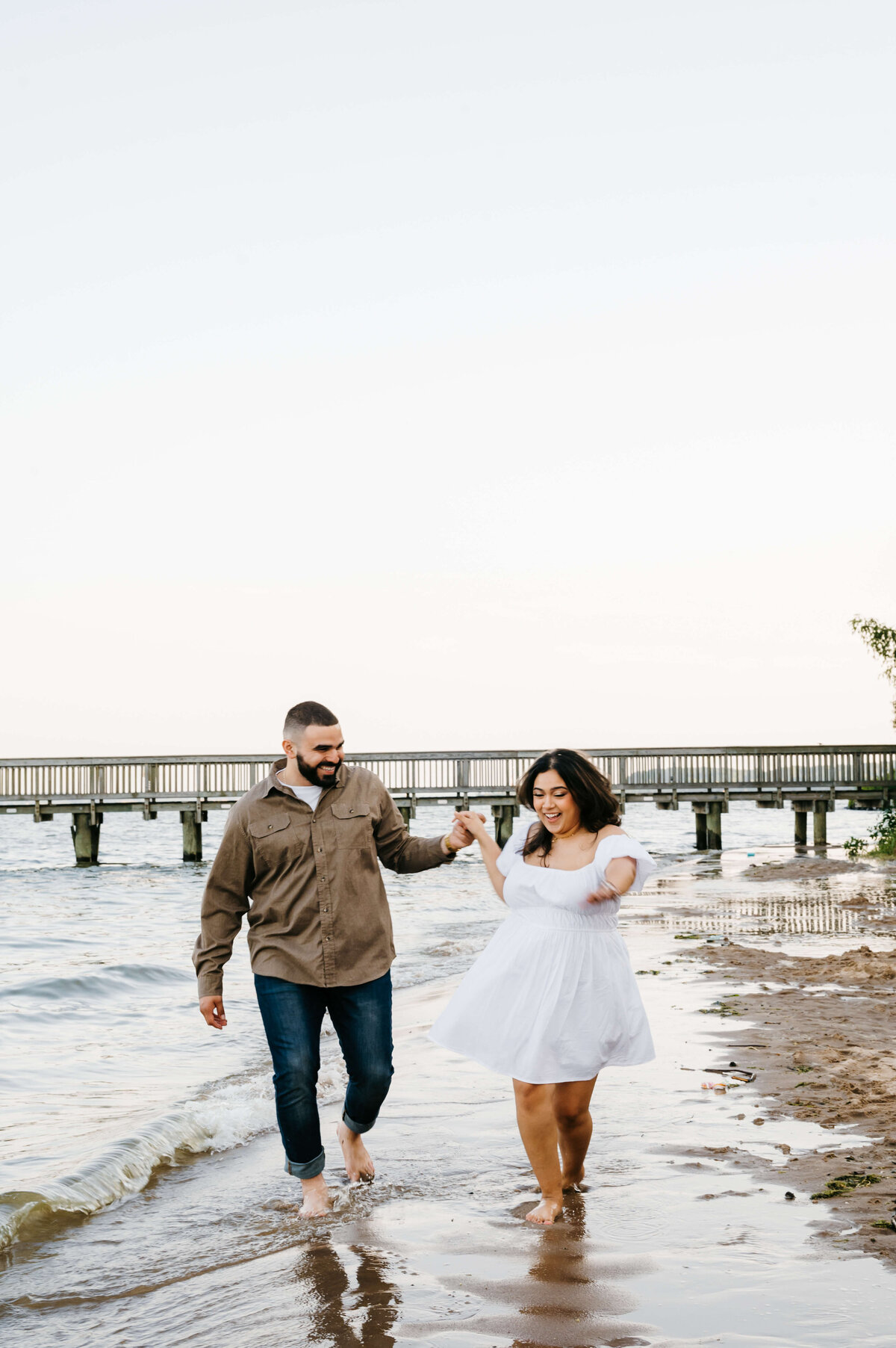 Virginia wedding photographer captures man and woman holding hands and running down the shore of woodbridge lake with a bridge in the distance while the couples laughs
