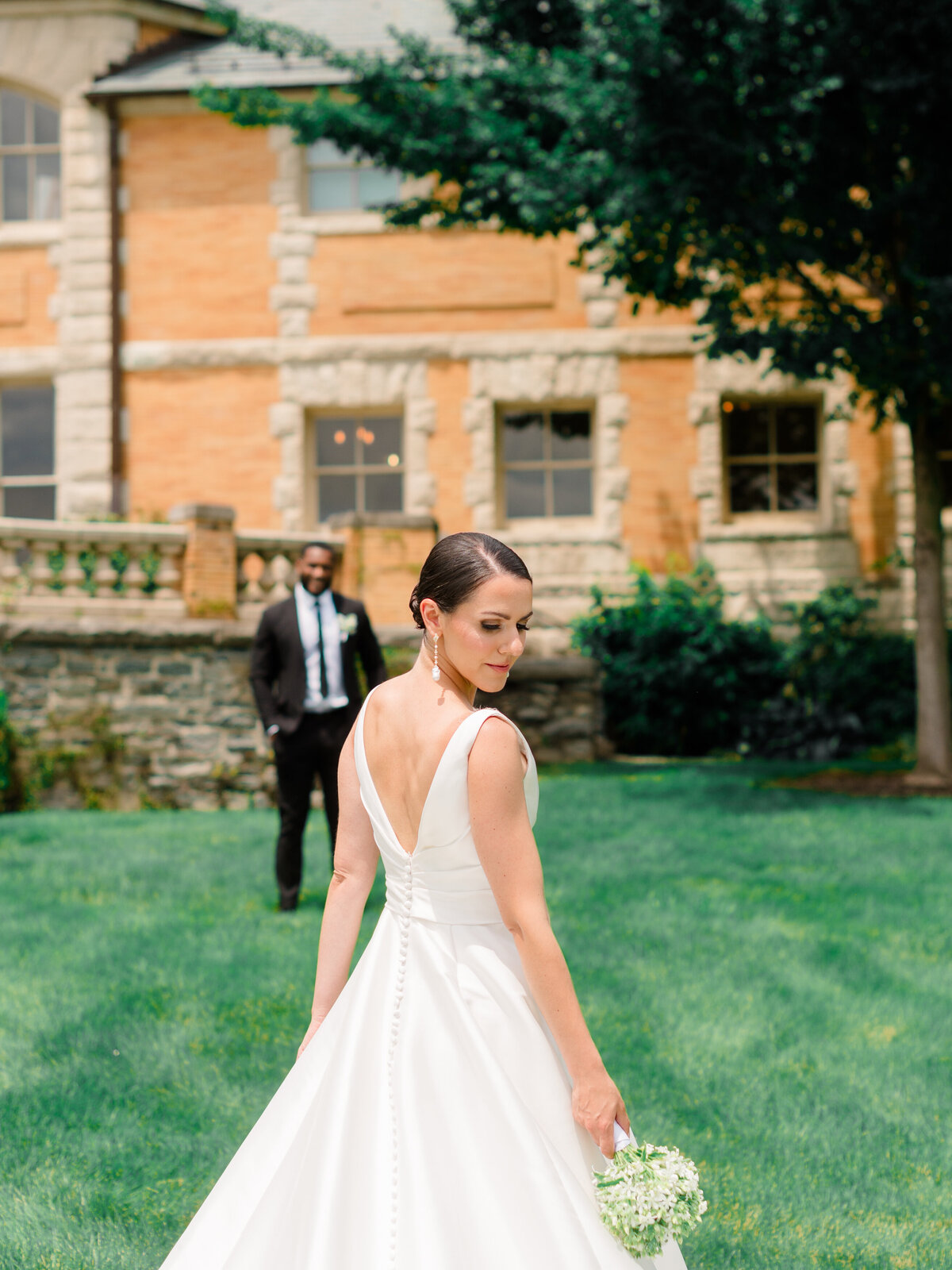 Elegant bride in a structured satin gown with a row of buttons down the back, photographed at Cairnwood Estate by Philadelphia Fine Art Wedding Photographer Elizabeth Kane. The image captures timeless bridal style with a romantic garden backdrop and a groom in the distance, evoking refined East Coast wedding charm.