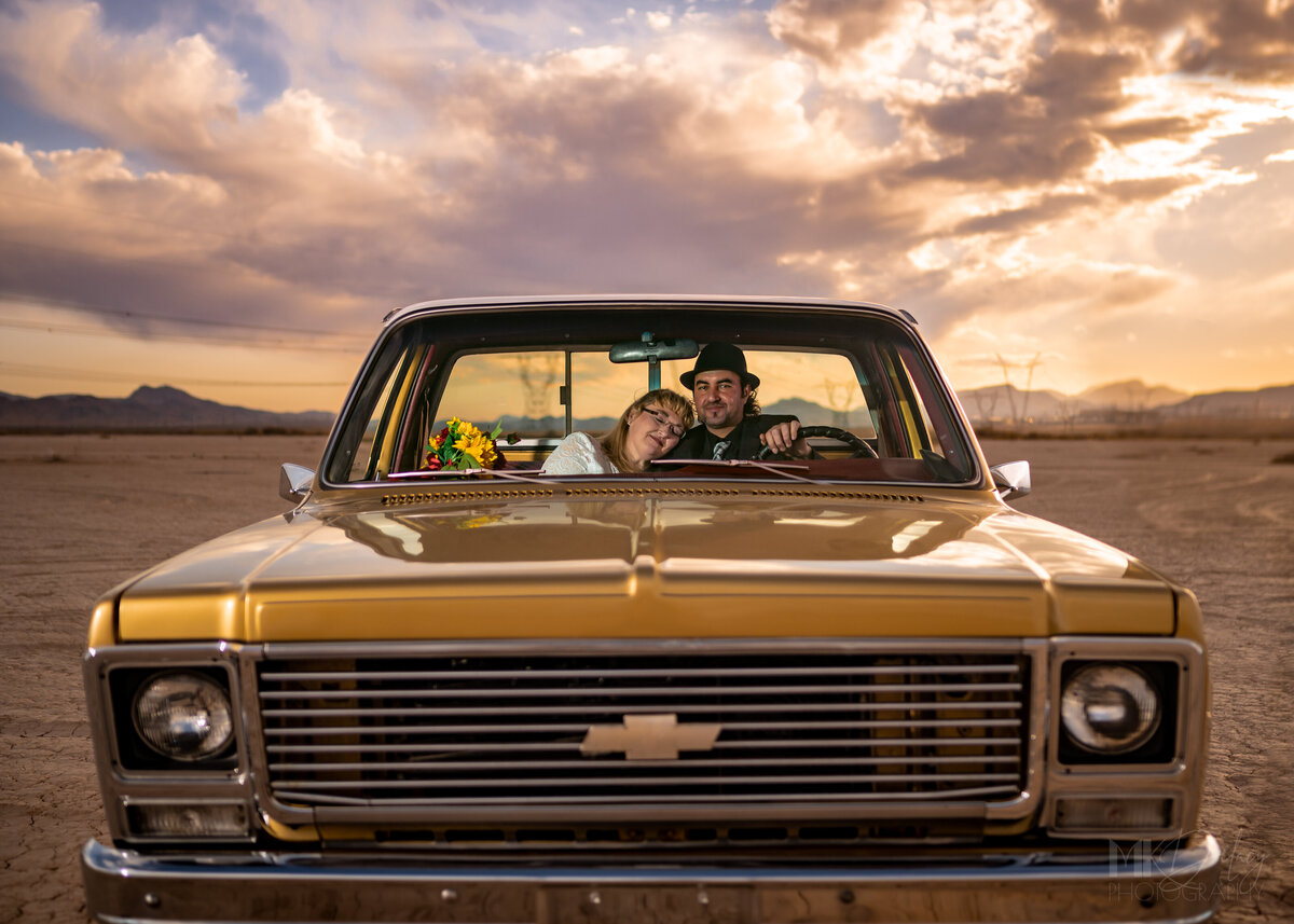 Bride and Groom in the Seat of his truck at golden hour with beautiful clouds with pink and purple skies  on a dry lake bed  Dry lake bed elopement bride in lace wedding gown with glasses groom in black suit white dress shirt with  fedora  classic chevy truck gold  restored with golden hour sunlight las vegas wedding photography las vegas elopement las vegas wedding photographers mk delacy photography