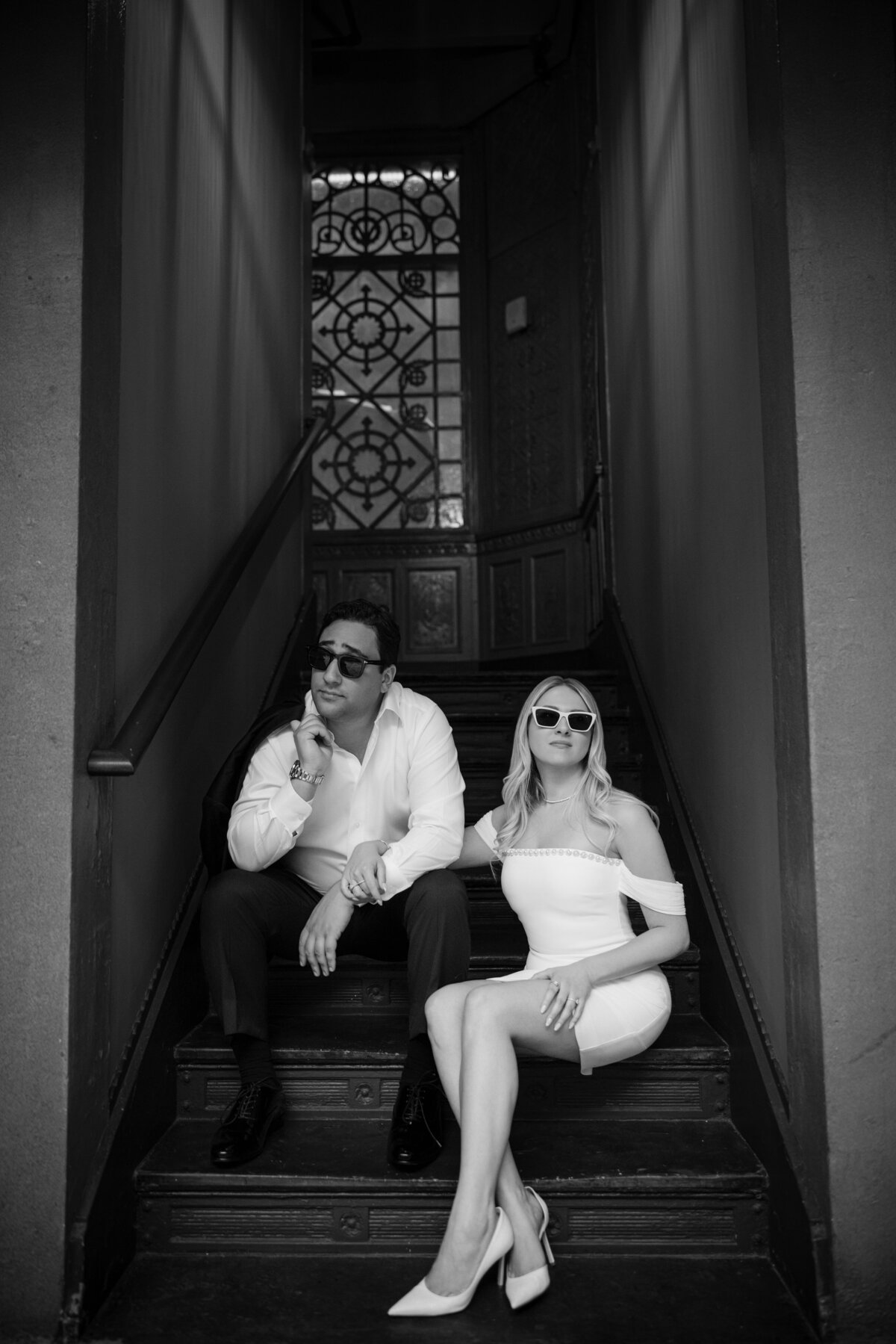 Black and white portrait of a stylish couple seated on the stairwell inside the Beekman Hotel, both wearing sunglasses and holding hands, framed by ornate ironwork and vintage tile—this image showcases the editorial potential of the space and underscores the value of securing a Beekman Hotel engagement photo permit for access to its dramatic interiors.