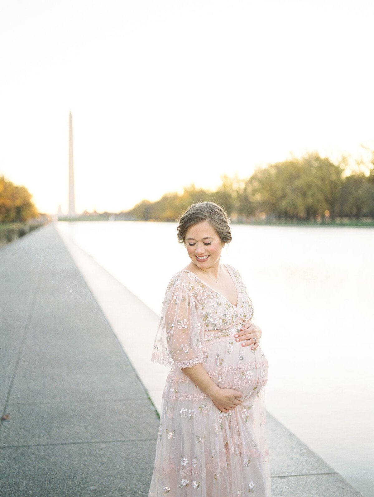 Pregnant mother stands with one hand on top of her belly, one hand below by Reflecting Pool in Washington, D.C.
