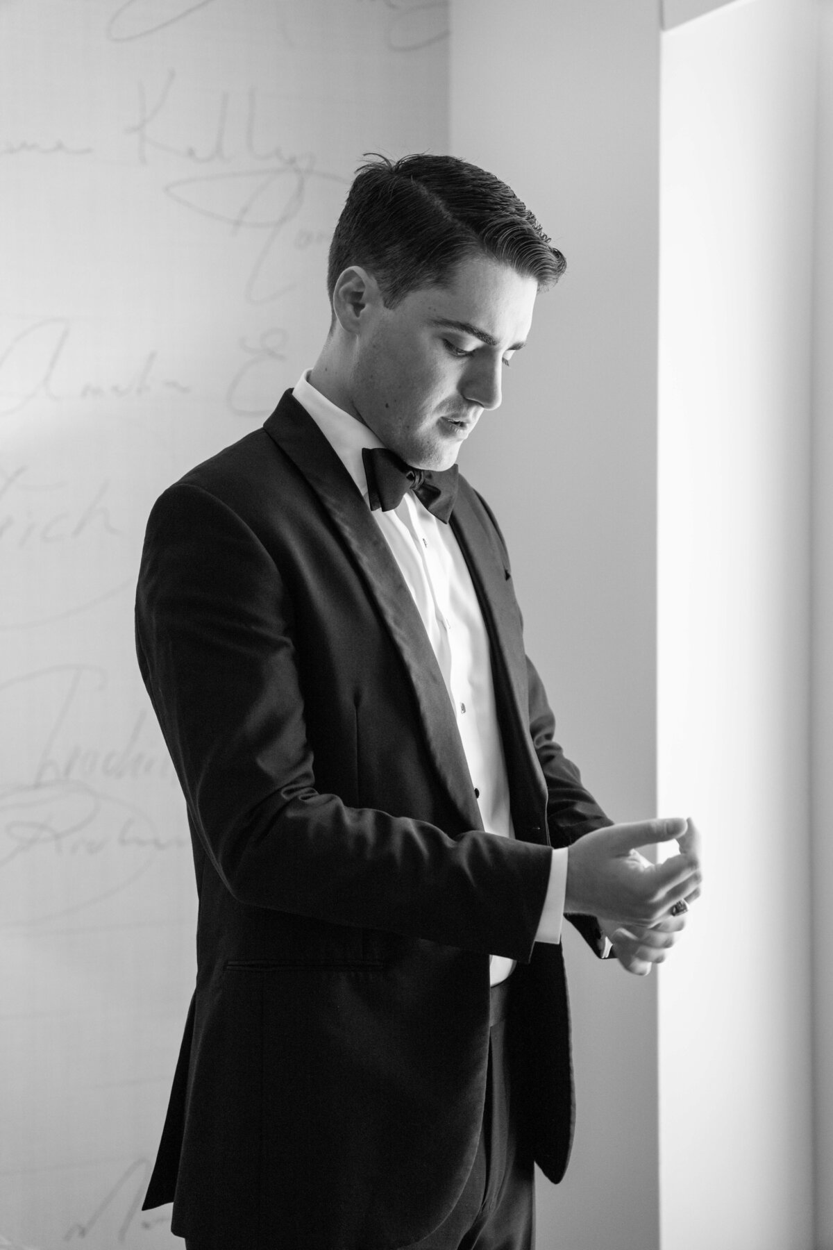 A man in a tuxedo adjusts his cufflinks in a softly lit room. He stands near a window, looking down with a focused expression. The background features a wall with faint signatures. The image is in black and white.
