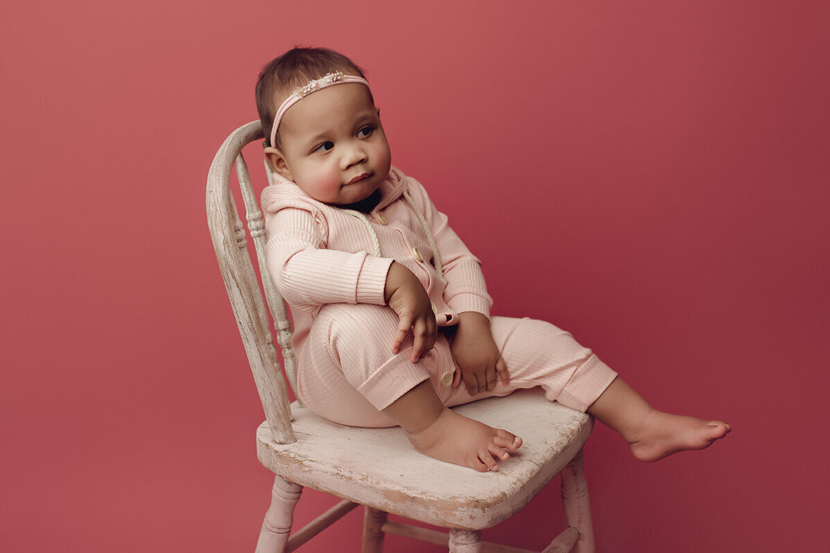 a one year old girl sitting on a white chair with her knee up and her hand resting on it