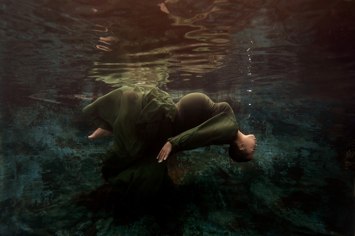 Underwater maternity photo featuring a woman elegantly arching backward in a flowing dark green gown, her body gently curving against a textured blue-green backdrop. The peaceful underwater environment highlights the artistry of this Dallas underwater maternity photographer, capturing the grace and tranquility of the moment