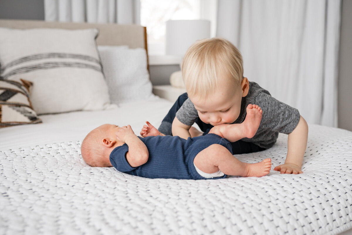 A toddler leans over to kiss a newborn baby on the foot, both lying on a bed with cozy white bedding.