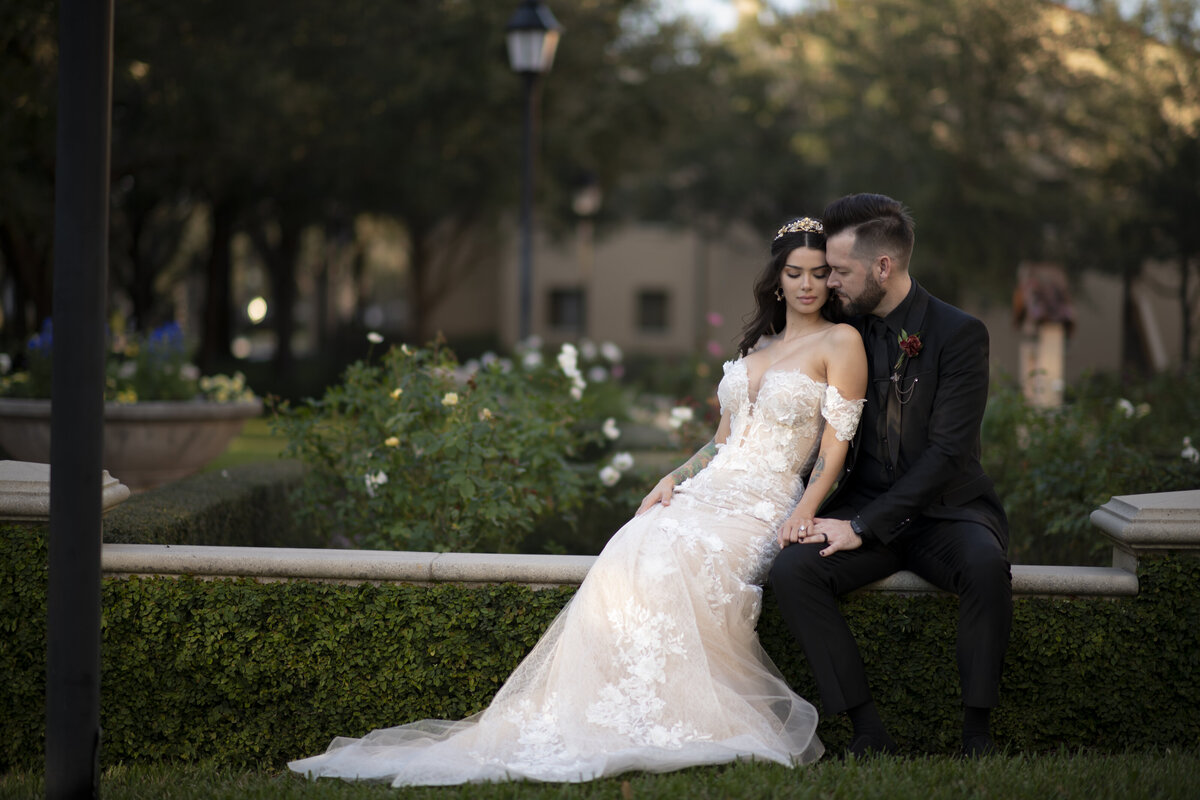 A bride in an off-the-shoulder lace gown sits with her groom in a lush garden, capturing a timeless and romantic wedding moment.
