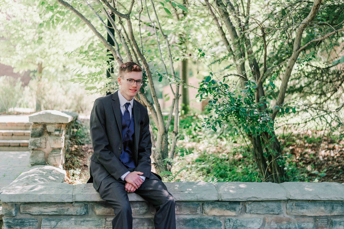 A handsome young man wearing a suit sitting on a low stone wall.