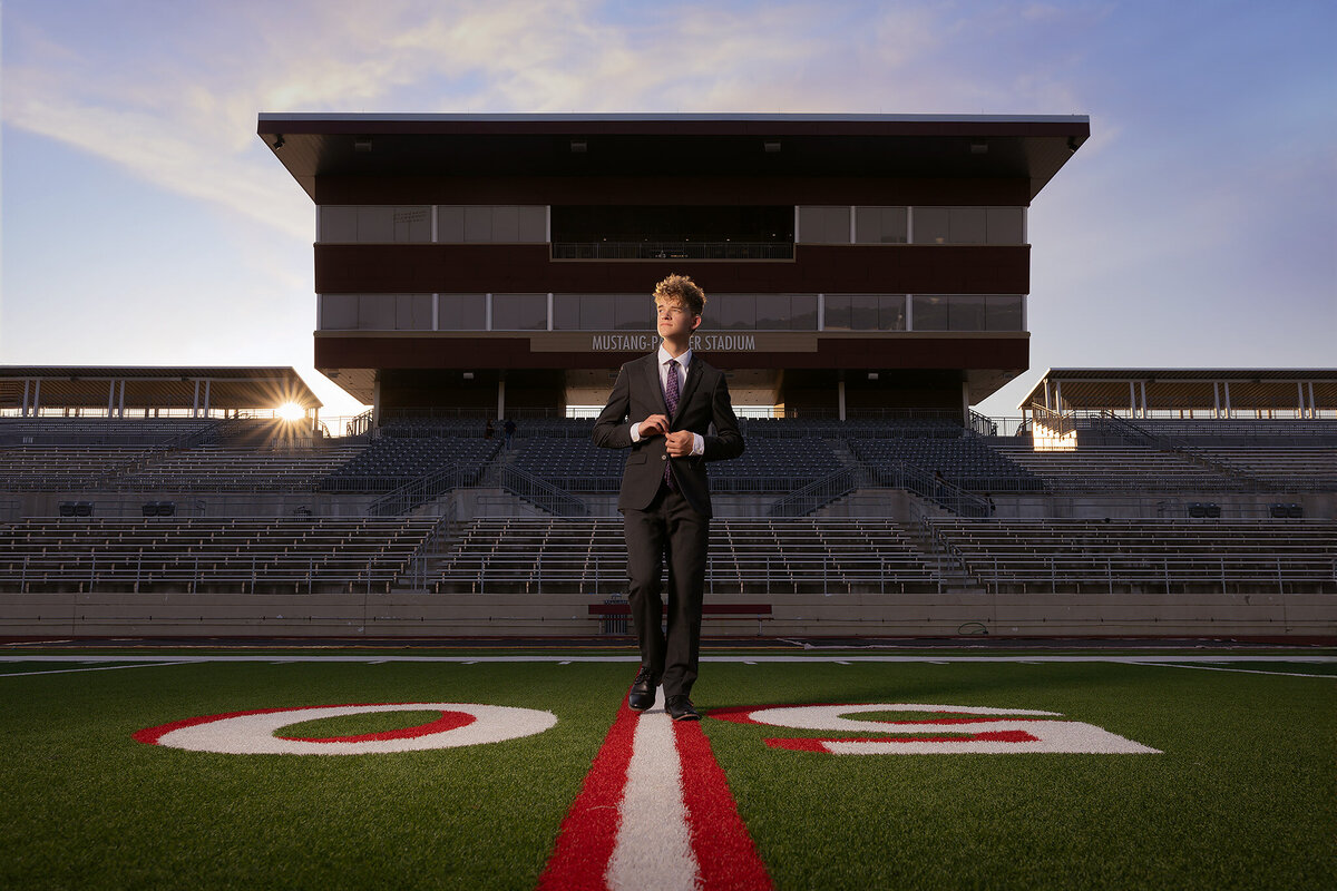 A high school senior adjusts his suit on the football field at Mustang Panther Stadium, captured during golden hour. The stadium stands in the background, and the senior is walking along the 50-yard line, embodying a confident, stylish look in this Dallas senior photography session.