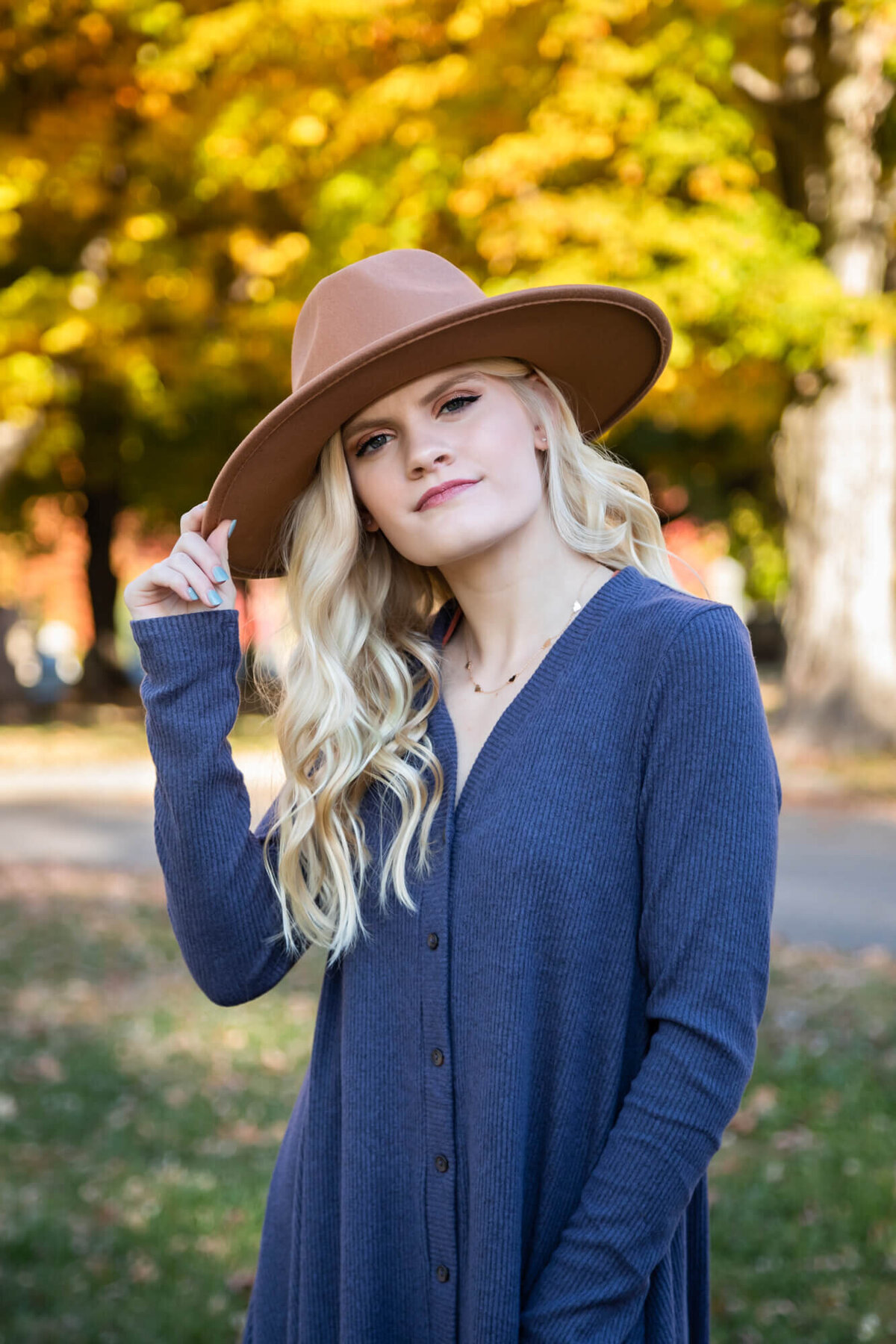 A lovely blonde teen posing for a senior portrait wearing a blue dress and a wide brimmed brown hat surrounded by golden Fall leaves. Captured by Springfield, MO senior photographer Dynae Levingston.