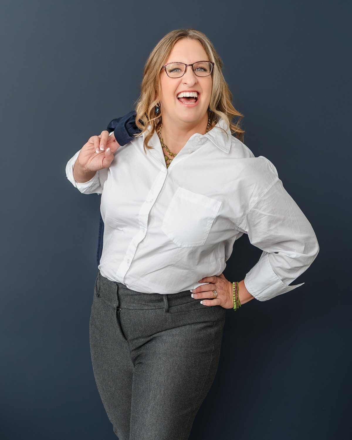 professional woman laughing at camera wearing white dress shirt and holding her suit jacket over shoulder against blue wall for headshot
