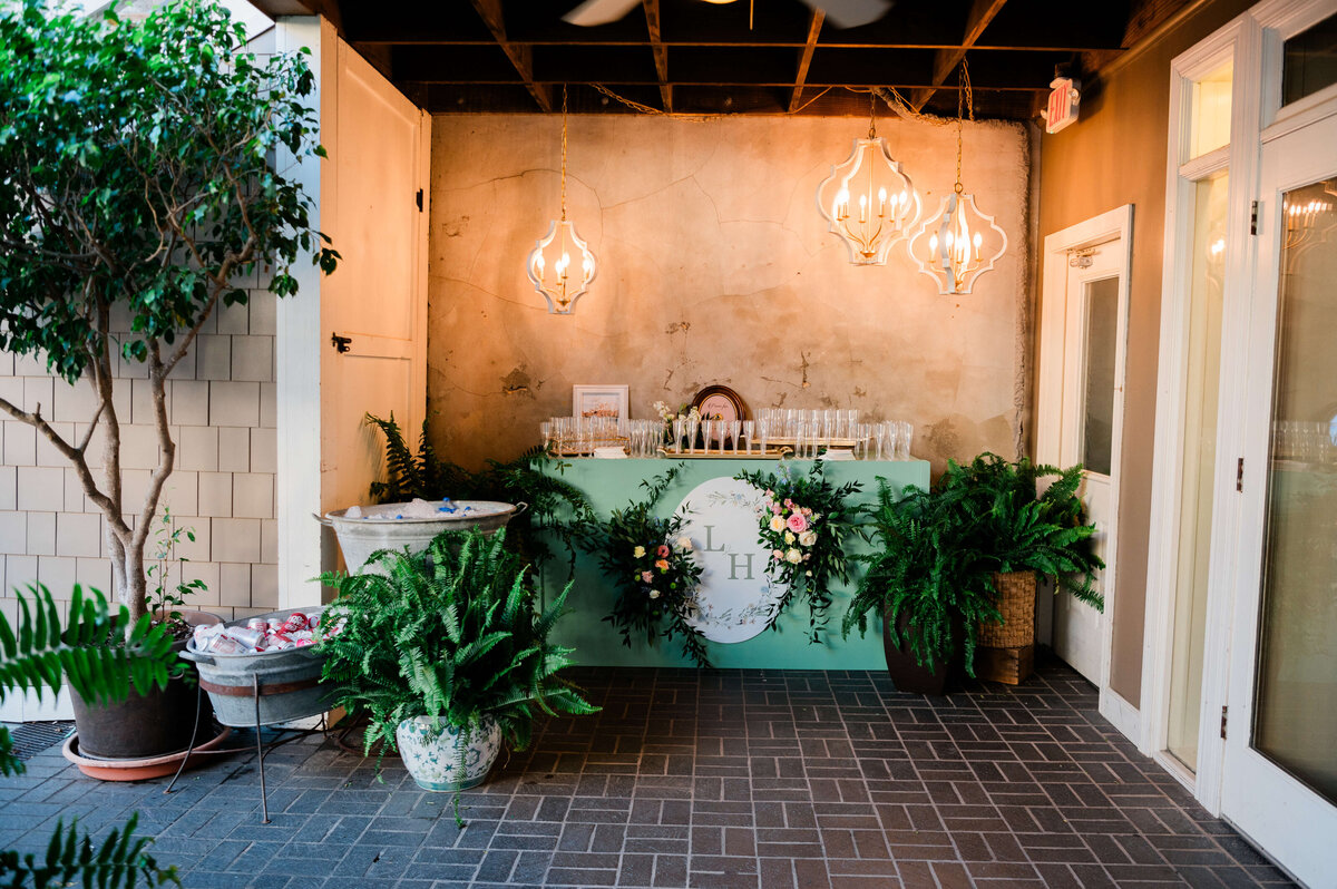 A green table decorated with a floral arrangement and ferns holds numerous filled champagne flutes. Behind the glasses is a round clock against a textured, beige wall.