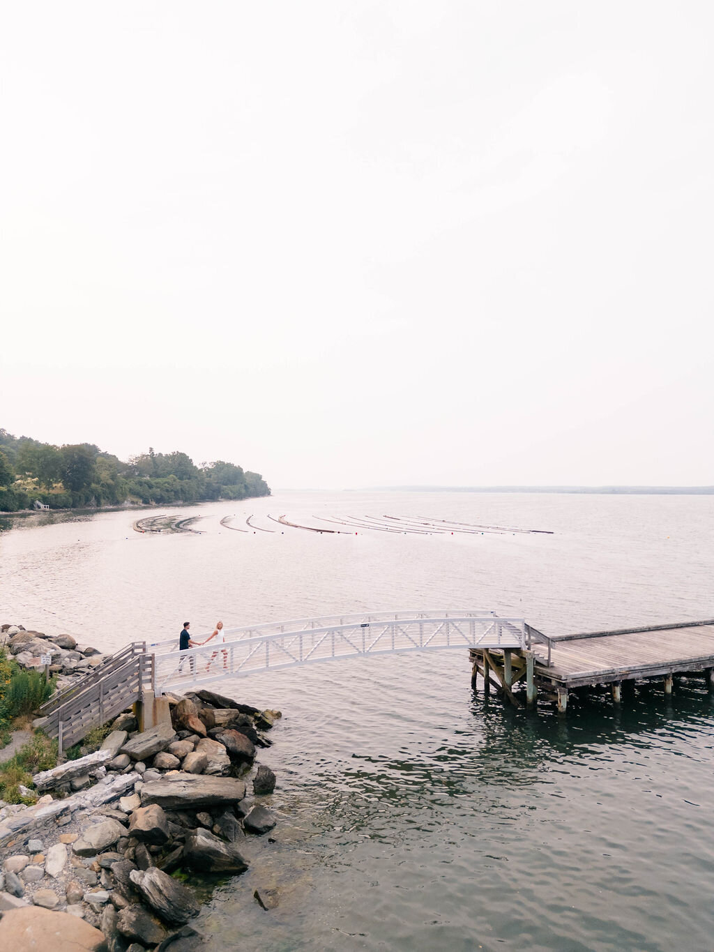 Wedding Photographer RI | A serene lakeside scene with two people walking on a wooden pier over calm water. The distant shoreline is lined with trees.