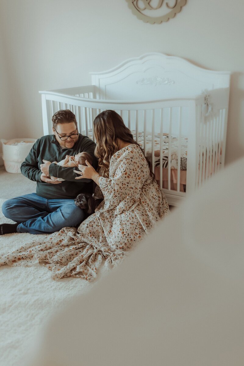 A couple sits on the floor in a nursery, holding a baby near a white crib. They appear engaged and content. A small dog is beside them.