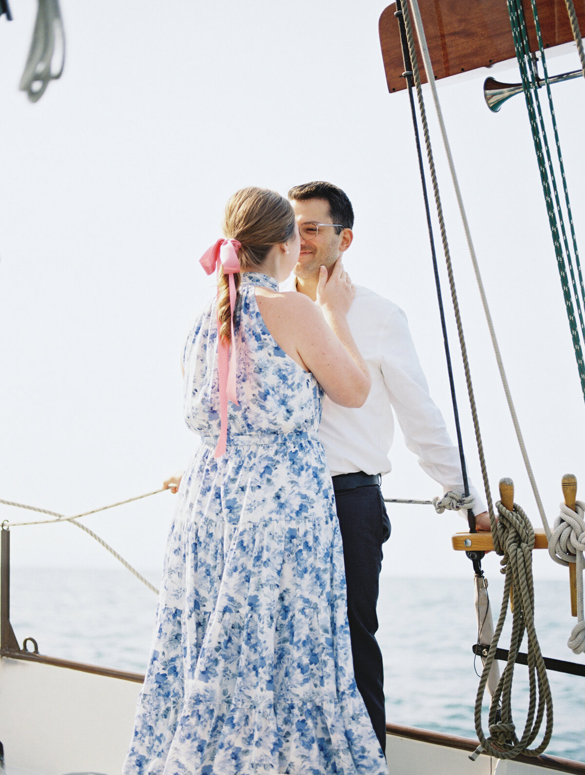 Engaged couple standing on a sailboat deck, gazing at each other, surrounded by ropes and sails.