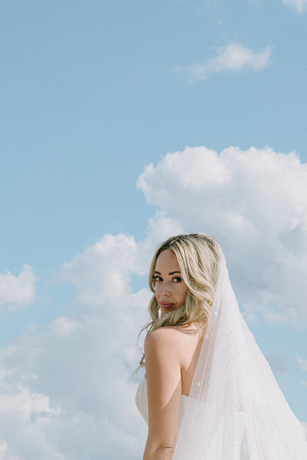 A stunning bride poses beneath a bright Texas sky with soft waves and glowing makeup, showcasing her dreamy pearl-dotted veil. Perfect inspiration for brides searching for wedding makeup artists in Dallas, San Antonio or Austin, or booking mobile bridal hair and makeup on location. Styled by the Mistique Makeup wedding beauty team.