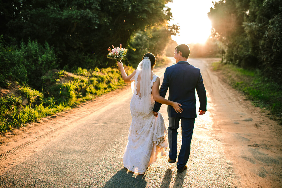 The bride and groom are dancing their way down a gravel road with the sun in the background. 