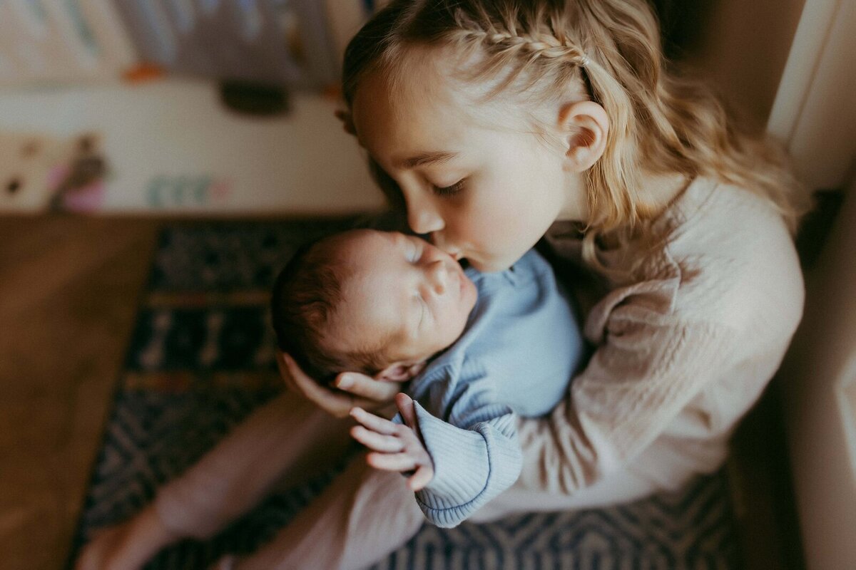A young girl with braided hair gently kisses a newborn baby on the forehead while holding them in her arms.