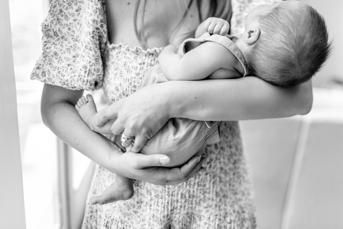 Close-up black and white image of a mother in a floral dress, swaying her newborn baby boy by a window during a quiet moment from a Kansas City in-home newborn session.