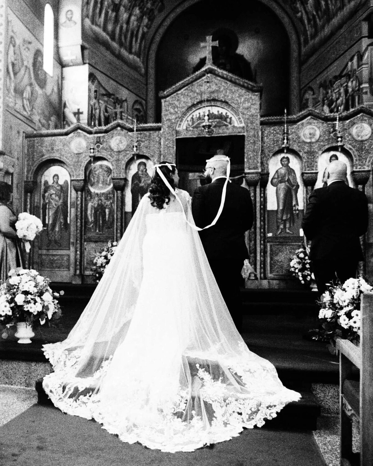 A bride and groom stand at the altar of an ornately decorated church during their wedding ceremony, facing the priest. Captured by a skilled NJ wedding photographer, the scene is surrounded by icons and elegant floral arrangements.