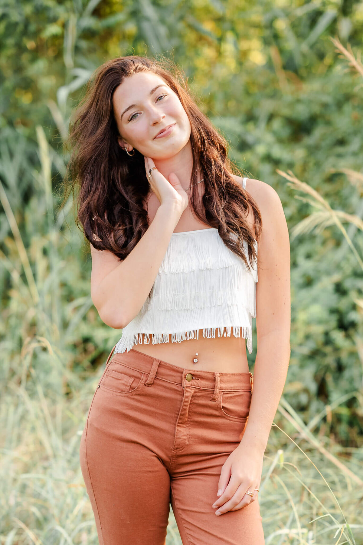 A high school senior, wearing orange pants and a white fringe top, smiles at the camera and puts her hand up to her face.