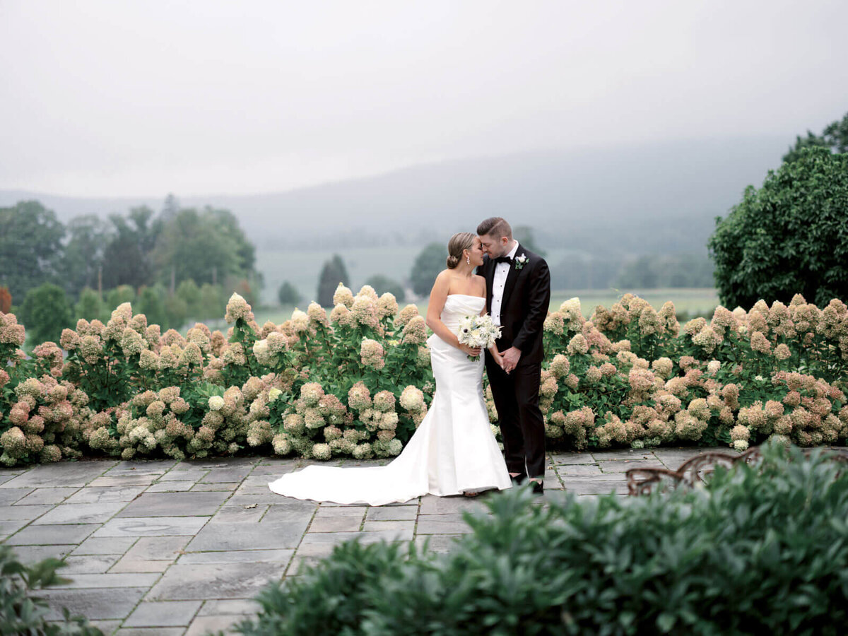 The bride and groom are romantically standing close, amidst beautiful flowers at Lion Rock Farm, CT.  Image by Jenny Fu Studio