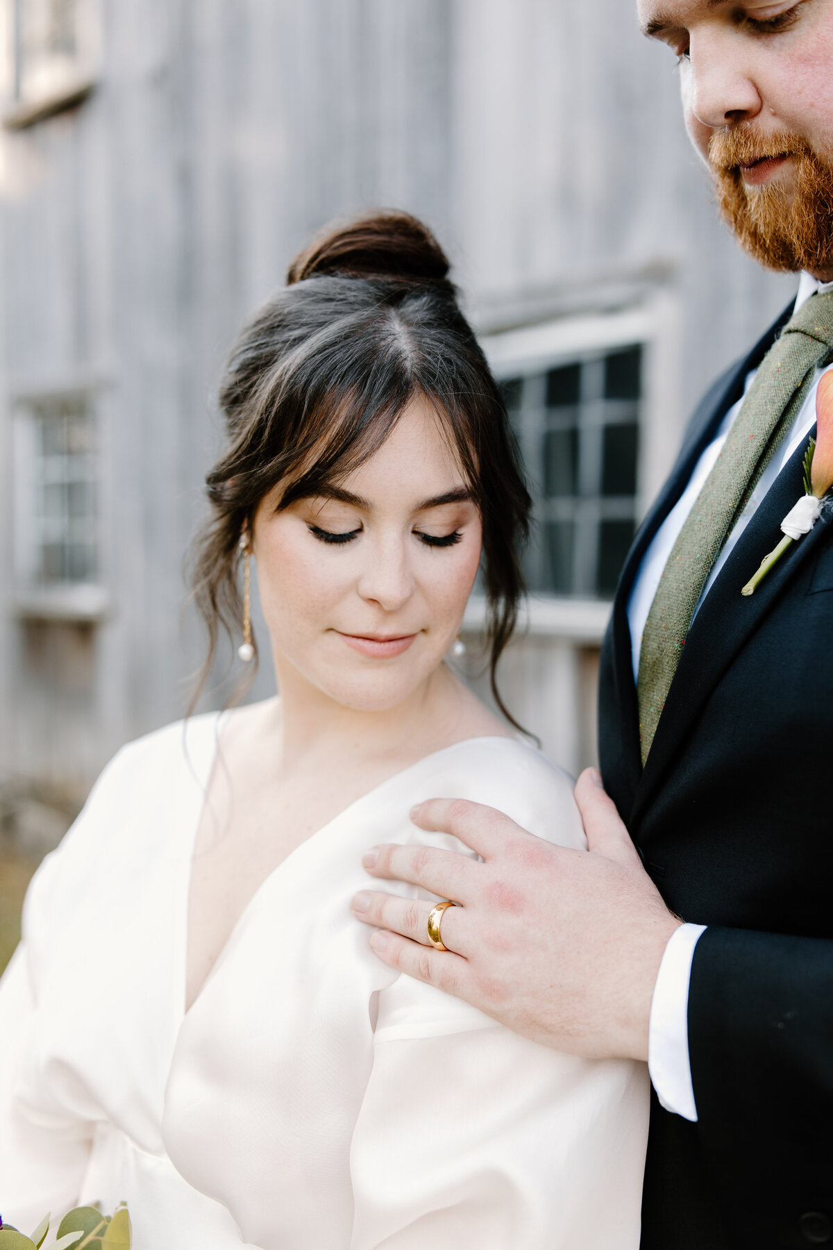 groom holding bride's shoulder displaying wedding band with both looking down