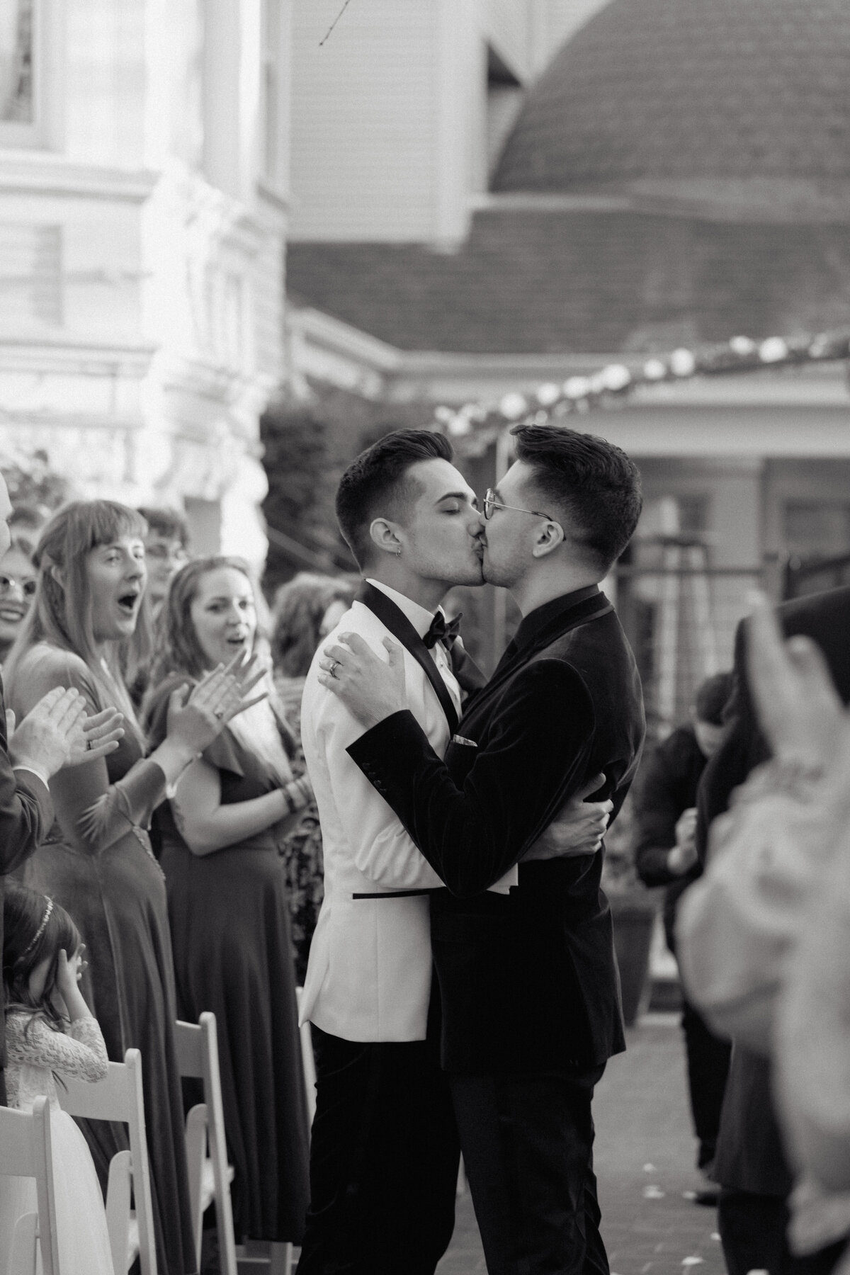 A newlywed couple shares their first kiss while guests cheer, captured in a candid and emotional cinematic wedding photo.