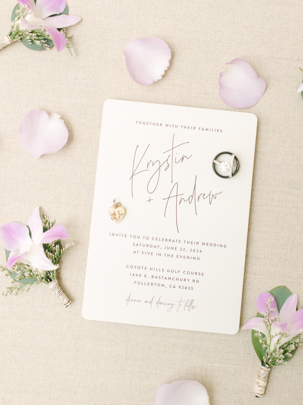 Elegant wedding invitation on beige fabric, adorned with pink flower petals and boutonnieres. The invitation features the names "Krystin + Andrew."