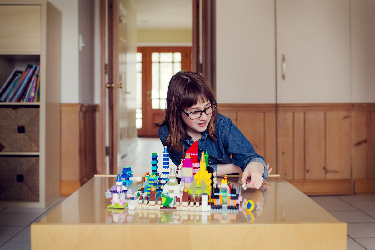 A girl is at a table  with her Lego creation in front of her, while adding to it.