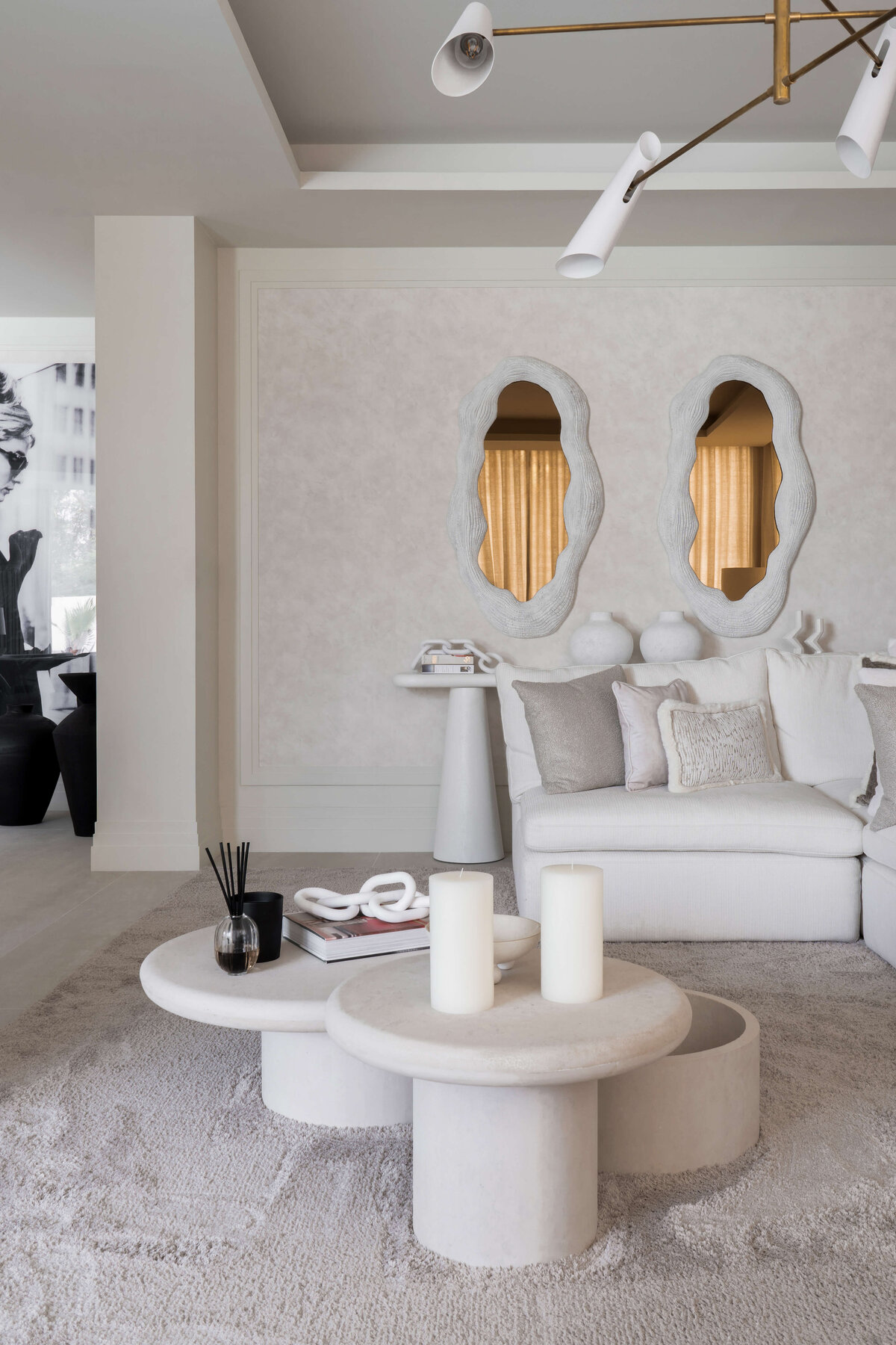 Spacious modern living room with a focus on light, neutral tones.  An interesting white center table with three levels. In the background, two contemporary white mirrors on the wall.