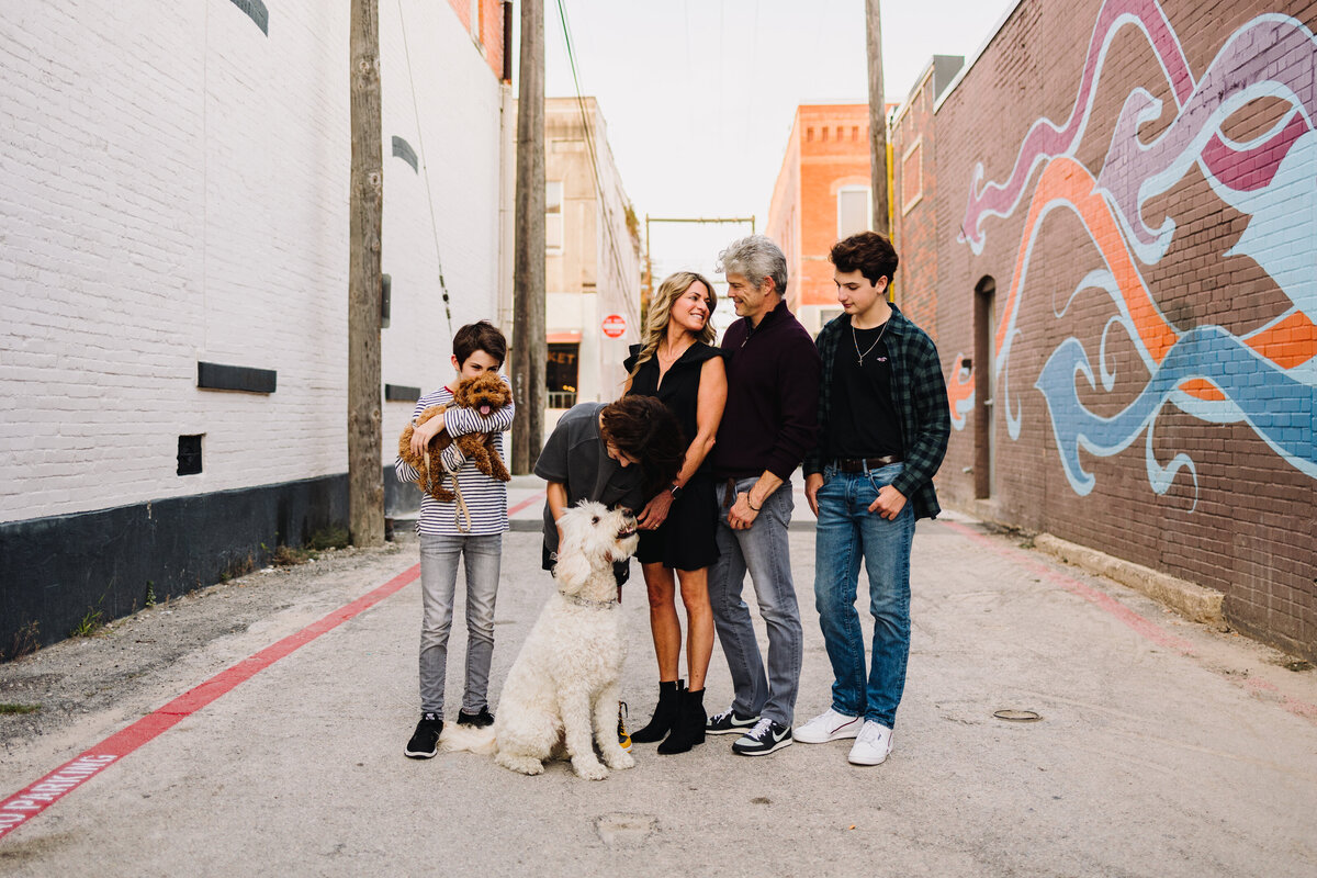 A modern family does a professional photo shoot. They are posing in an urban location on a wall with a colorful illustration. The woman is wearing a short black dress, the man is wearing jeans and a black T-shirt, and the children are playing with their dogs.
