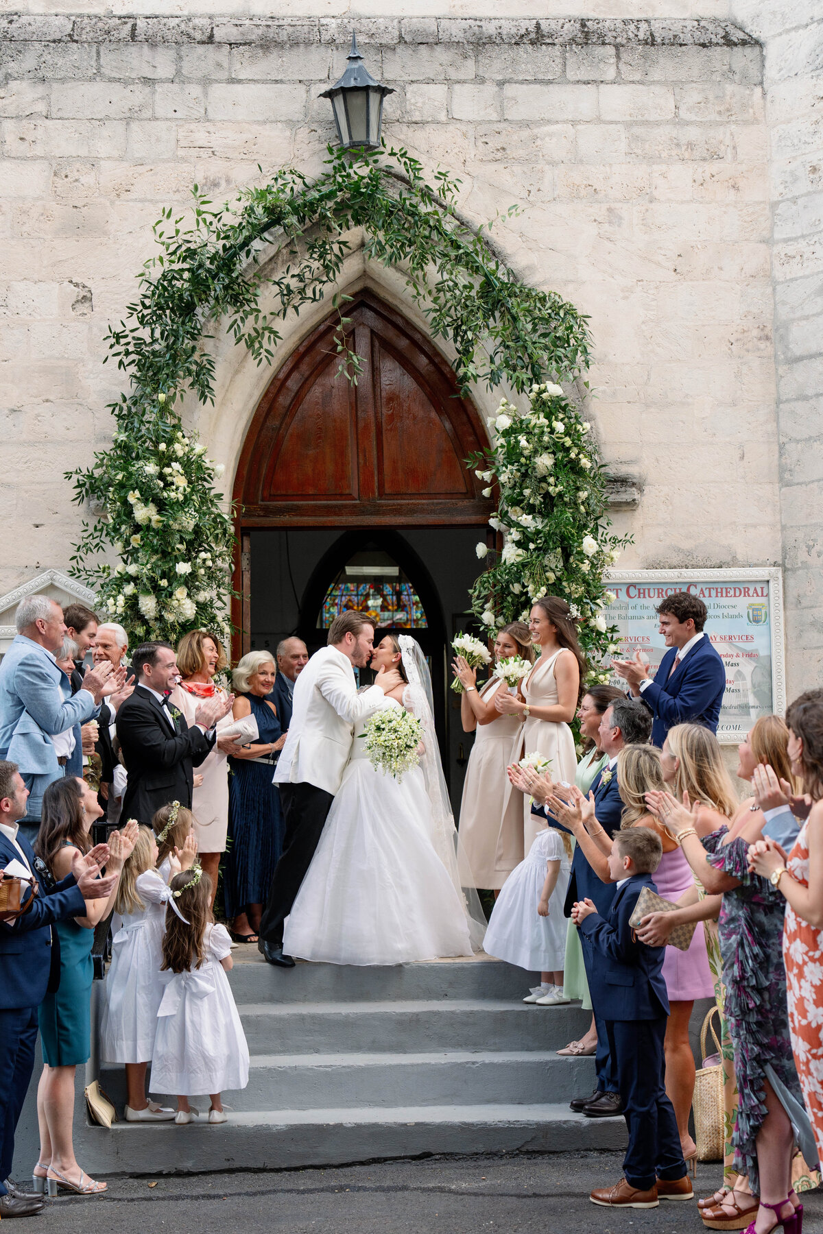 Bride and groom share a kiss on the steps of a historic stone church, surrounded by cheering guests and flower girls, after a romantic destination wedding, captured by Mari Harsan Studios.