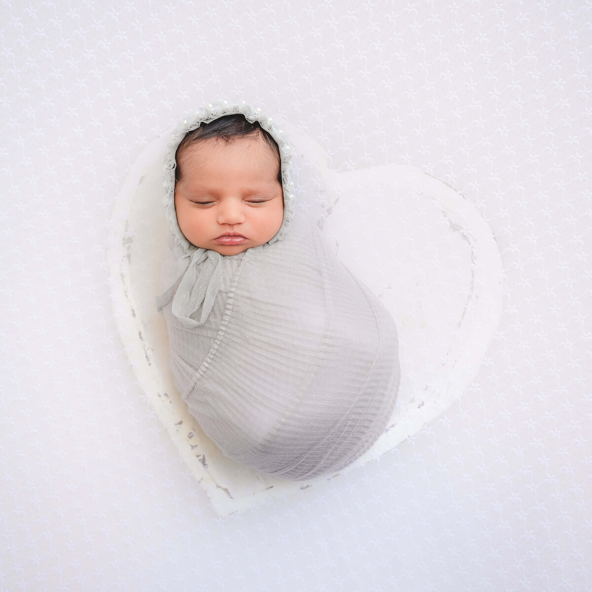 newborn girl swaddled in light gray wrap with matching bonnet.  She is posed sleeping in a white heart bowl. Image by E. Benecki Photography.