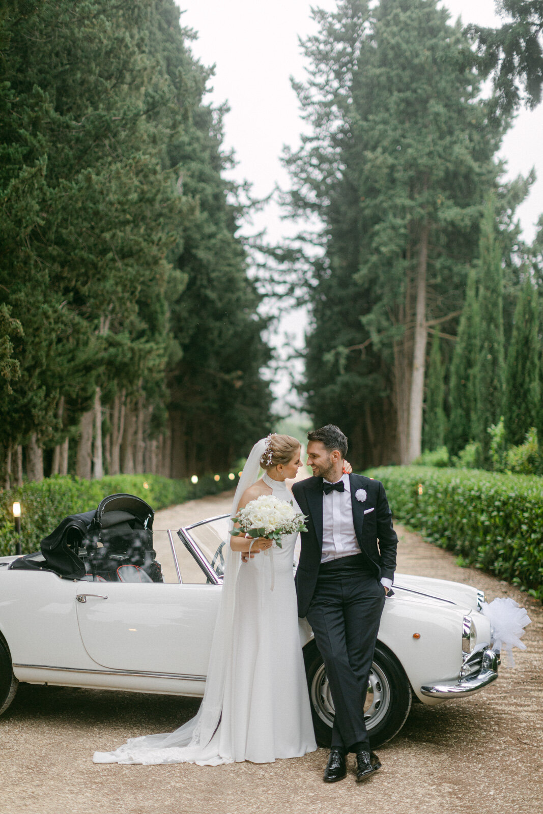 Bride and groom with vintage Italian car in Tuscany – romantic wedding portrait in a cypress-lined driveway.