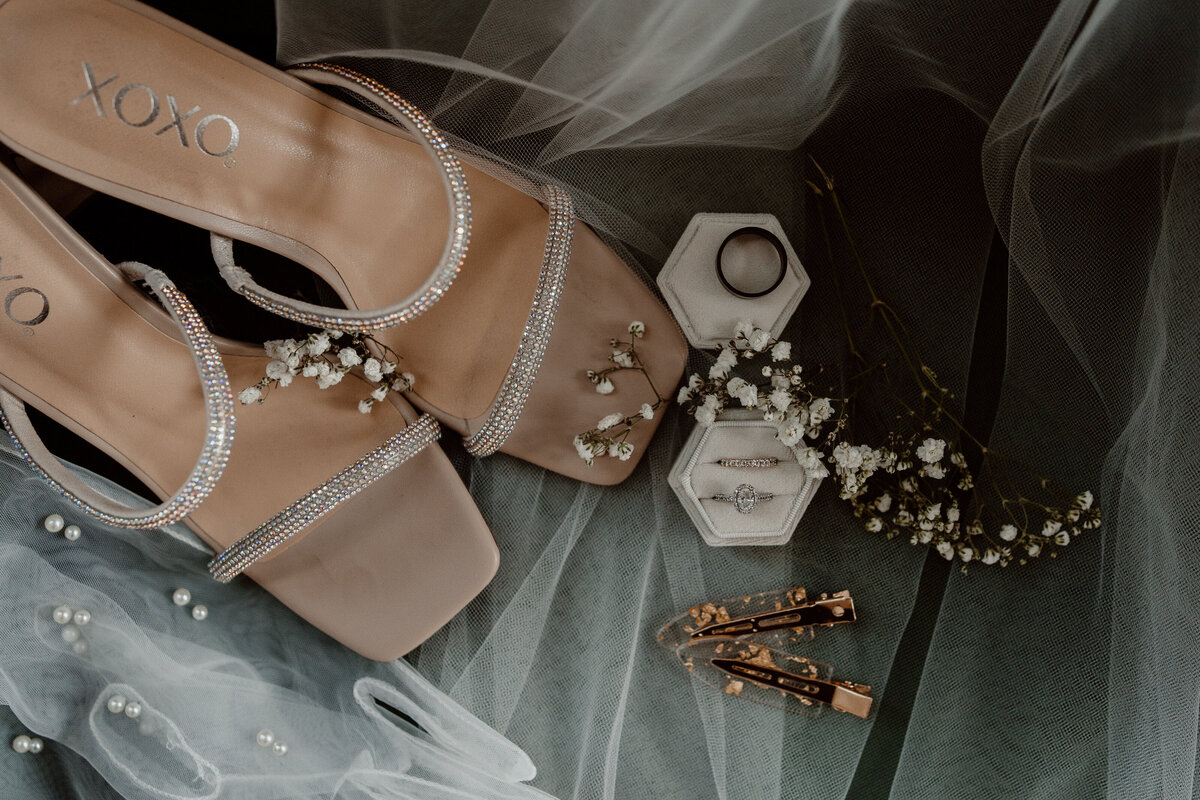 Elegant wedding detail shot featuring rhinestone-embellished heels, wedding rings in a hexagonal box, delicate baby's breath flowers, and hair accessories on a soft tulle background. Ideal for couples looking for a candid and documentary-style wedding photographer to capture stylish and intricate bridal elements.