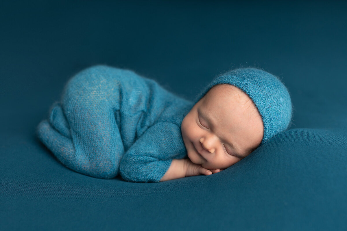 Newborn baby in a teal onesie curled up asleep on a seamless teal fabric – Keller newborn photography session.