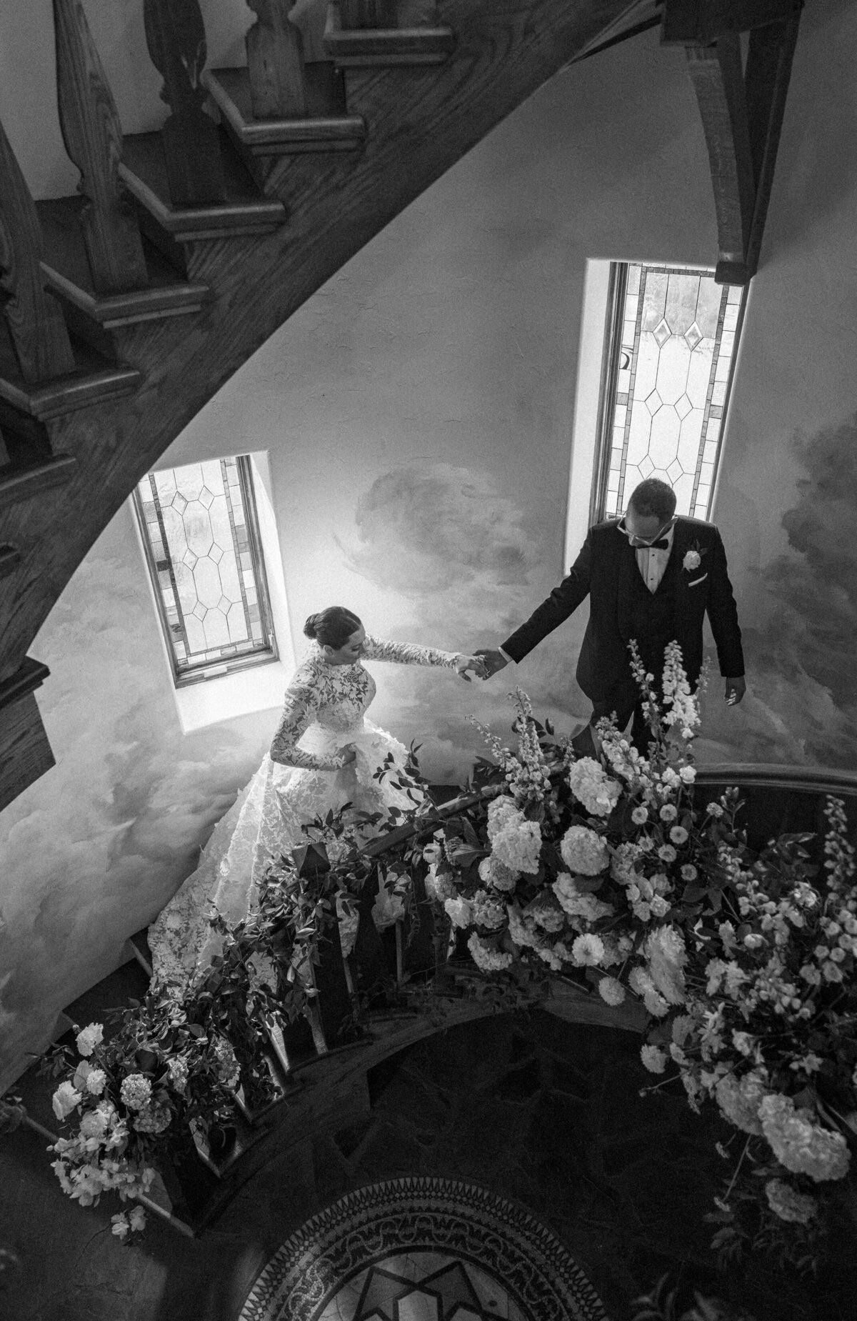 Elegant black and white image of bride and groom on a stone staircase surrounded by lush floral installations inside the castle.