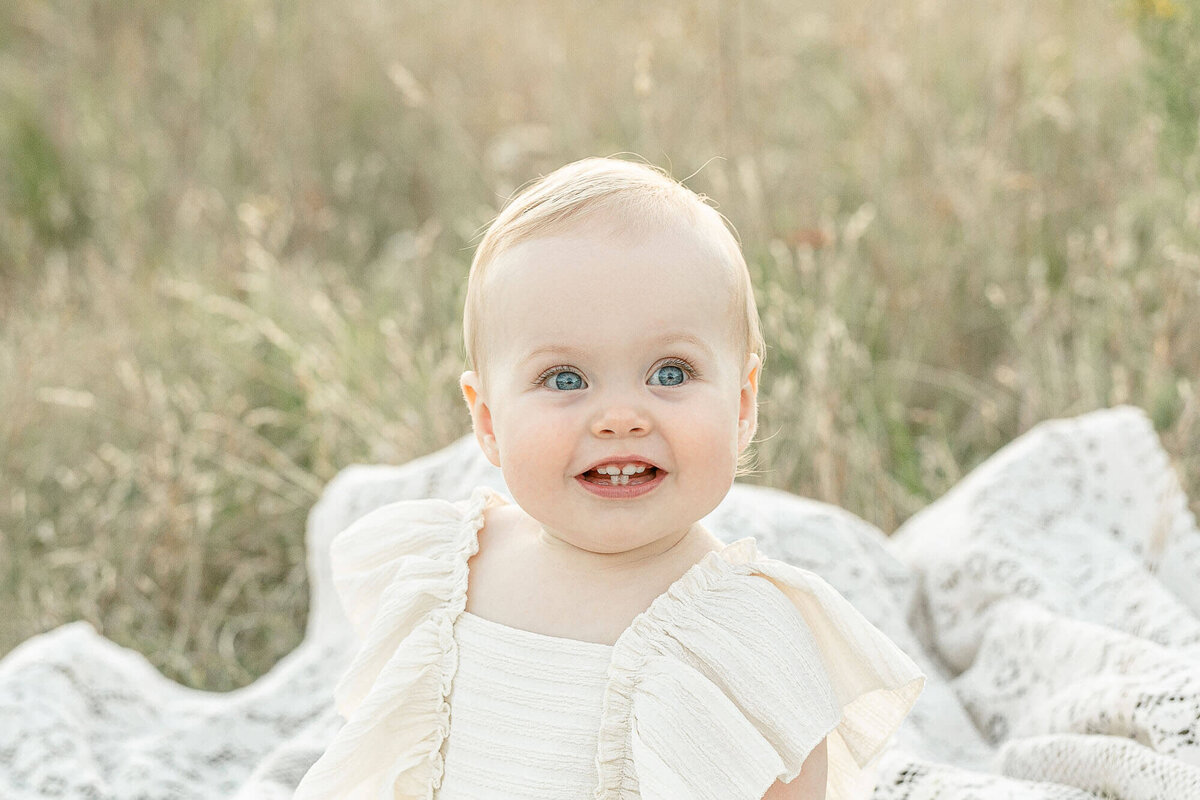 One year old baby girl in a cream dress is sitting on a cream lace blanket in a field of golden grasses at sunset. She has a big smile on her face and is looking a little off camera. She has very light blonde hair.