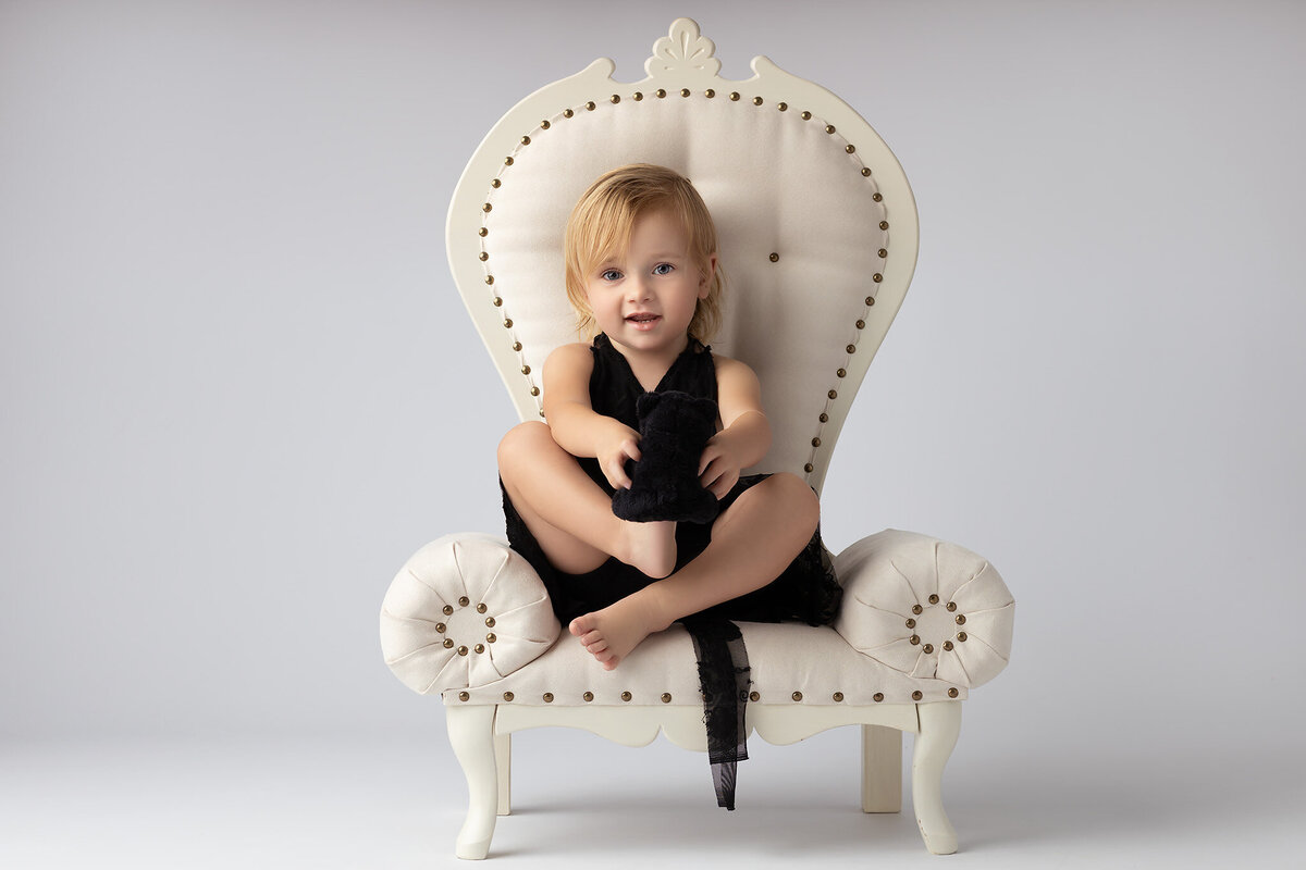 Toddler sitting on a white chair holding a black stuffed animal, captured by Dallas tx family photographer.