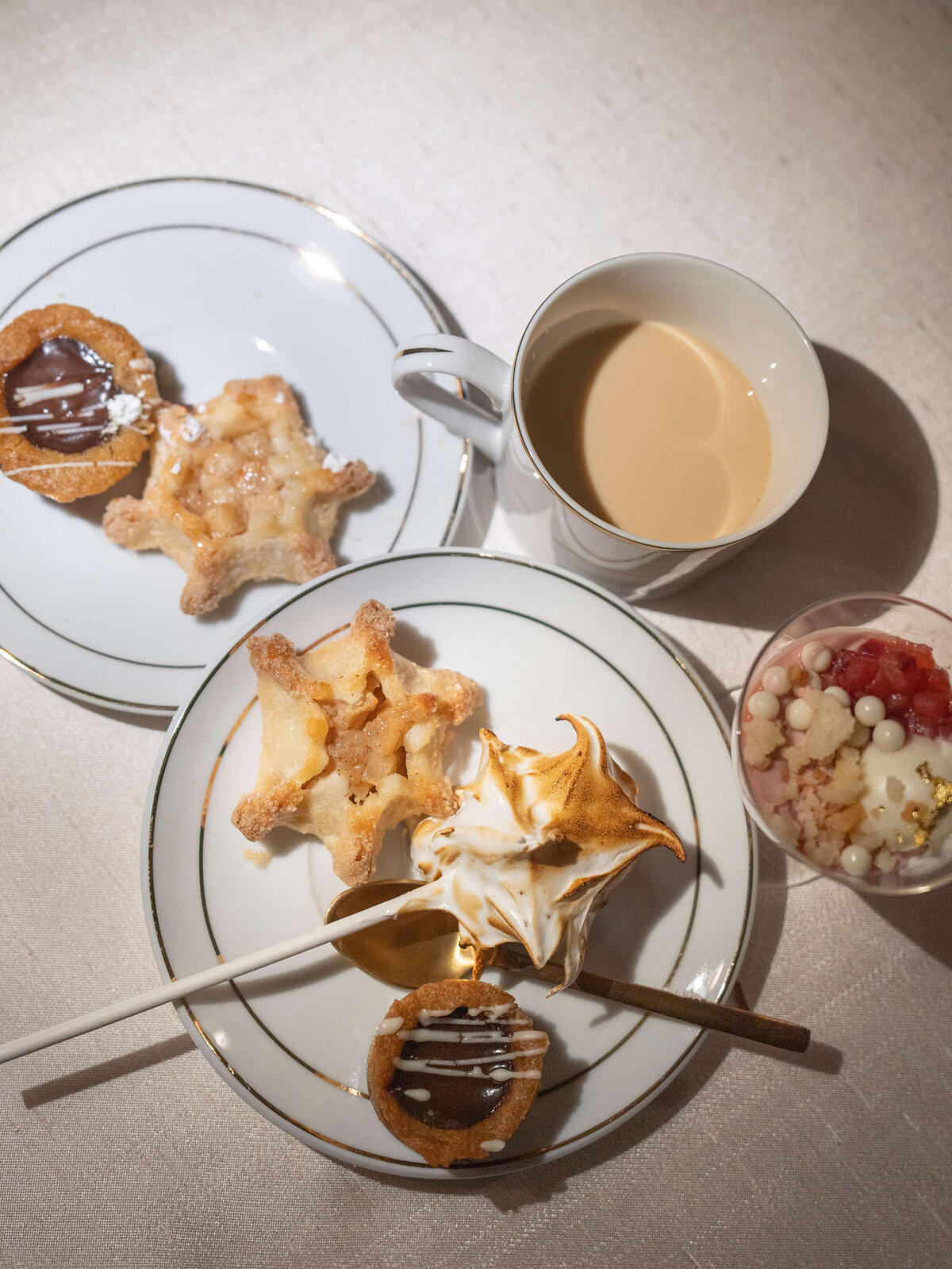 Two plates with pastries, one with star-shaped desserts and the other with a meringue-topped treat, accompanied by a mug of coffee and a cup with pink and white desserts. A spoon rests on the plates, creating a cozy table setting.
