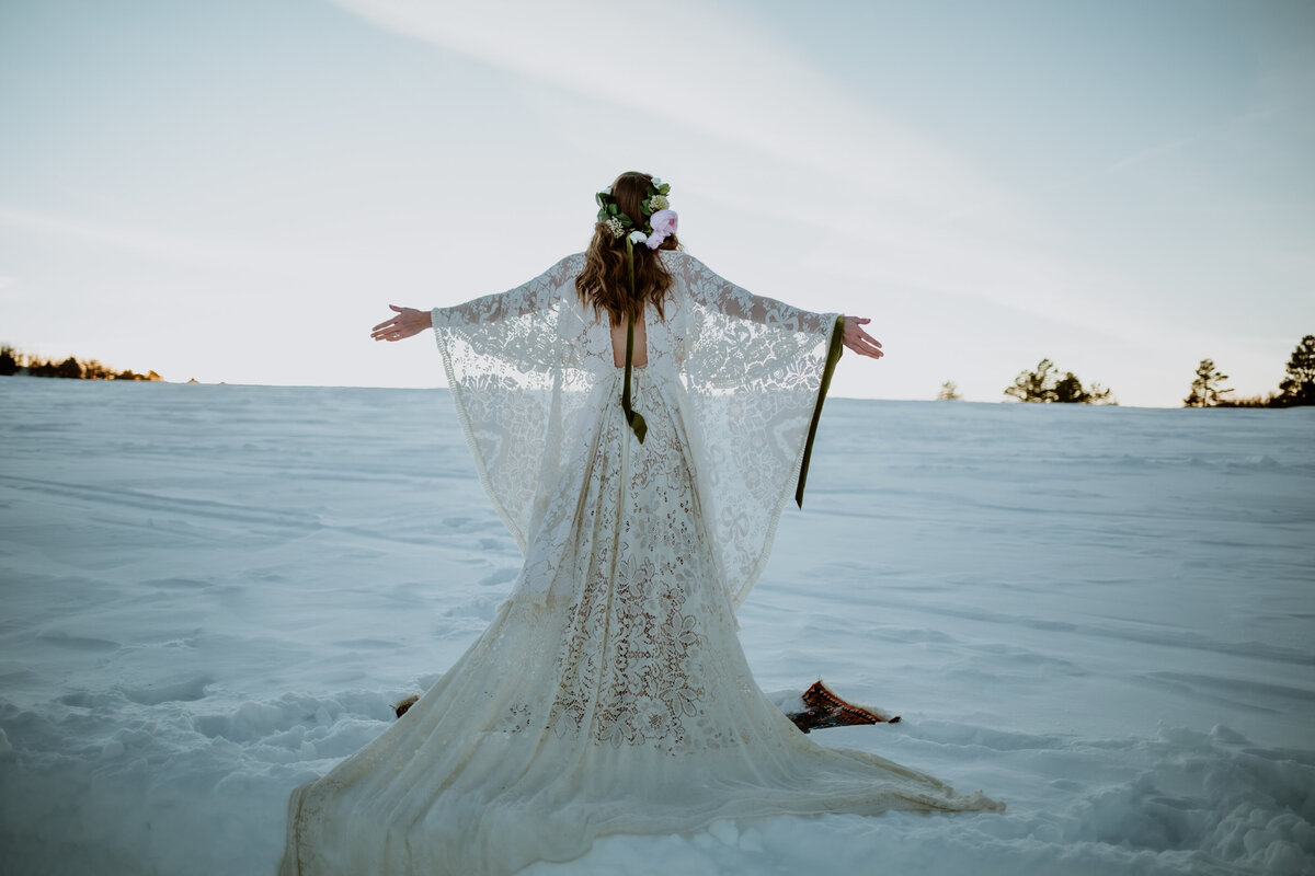 Bride stands in the snowy mountains of Ouray, Colorado with arms outstretched in a lace wedding gown