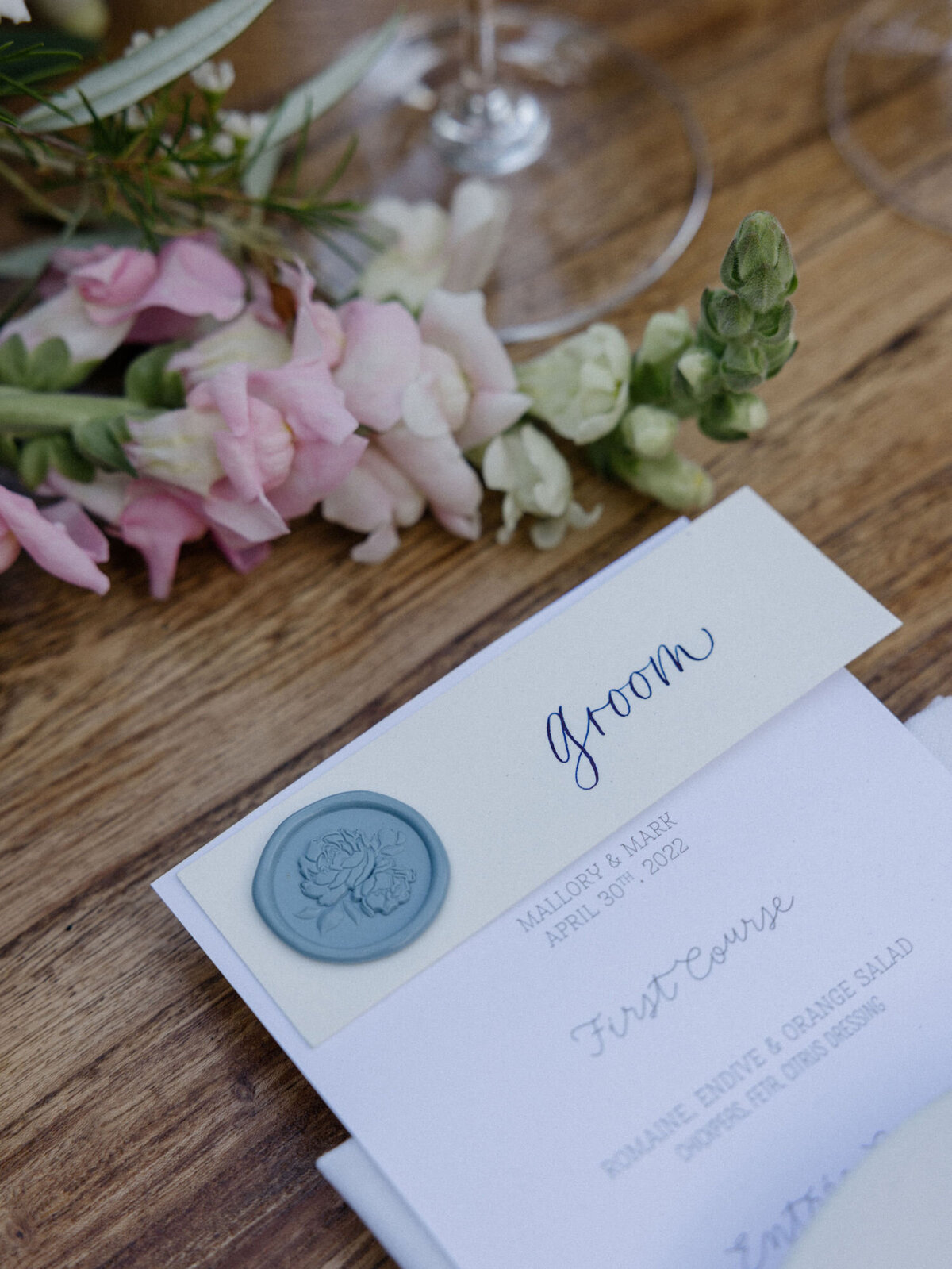 Calligraphy place cards with a dusty blue wax seal and navy lettering on top of a menu for a wedding tablescape