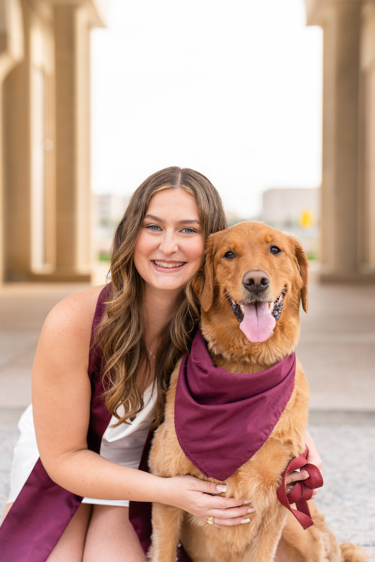 Texas A&M senior girl hugging dog while wearing white dress and Aggie stole in the archways of the Bell Tower