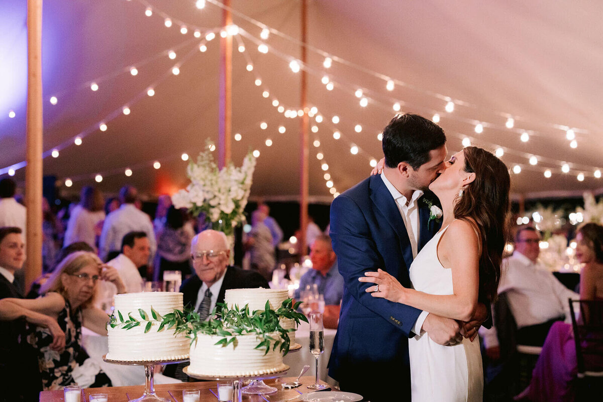 The bride and the groom are kissing each other beside the wedding cakes, as the guests watch on, in Cape Cod Summer Tent, MA.