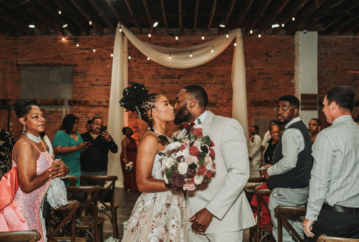 A beautiful couple shares a passionate kiss during their emotional wedding ceremony at the industrial-chic Neon House in Norfolk, Virginia, featuring exposed brick walls, string lights, and joyous guests, captured by Quinn Photography LLC.