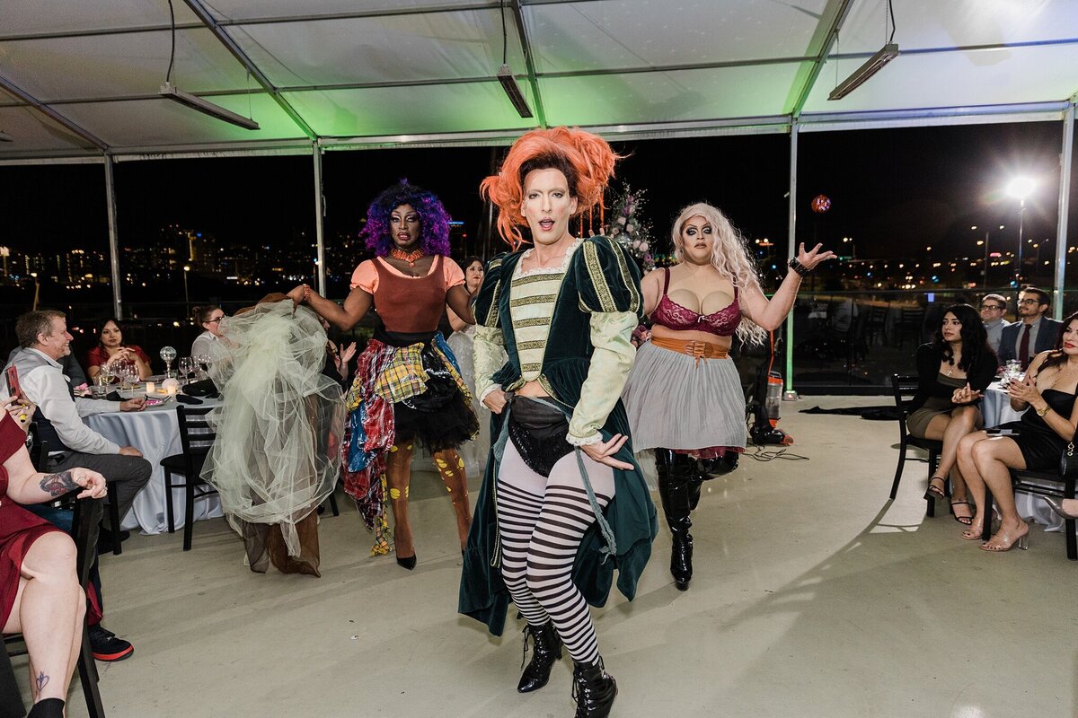 Action shot of three drag queens dressed up as characters from the three witches from the movie Hocus Pocus at a wedding reception in Dallas, Texas. Wedding guests cheer them on from their reception tables on either side.