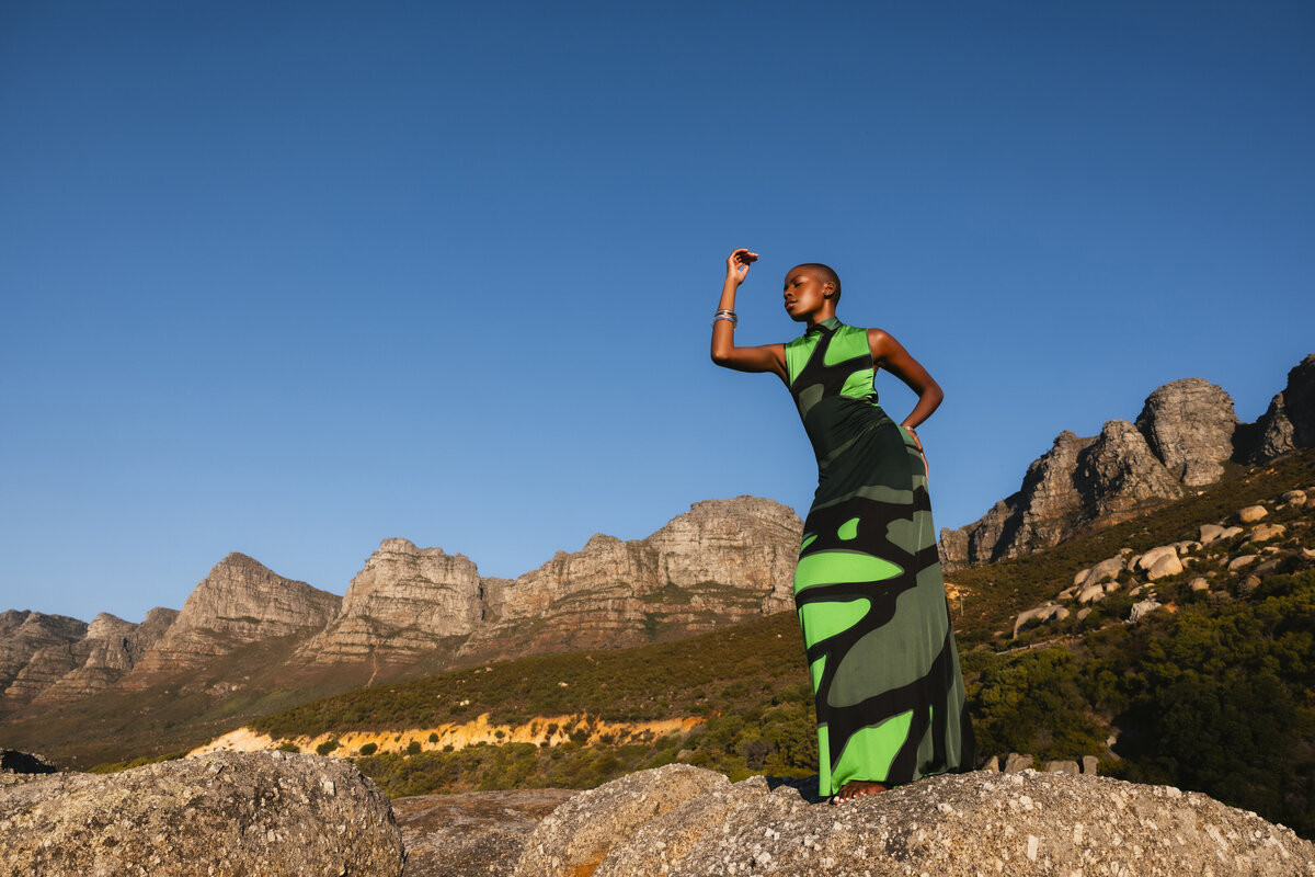 Model standing powerfully on a rocky cliff in a bold green and black dress, framed by the dramatic mountains of Cape Town and a vibrant blue sky, evoking strength and elegance.