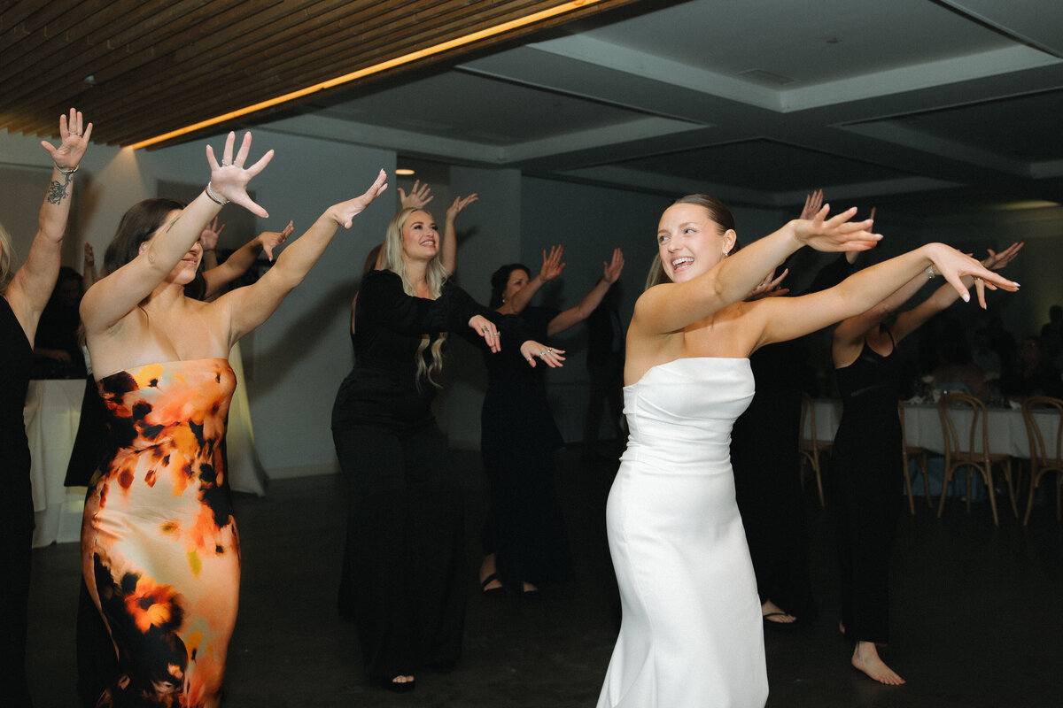 A bride in a white strapless, fitted wedding dress raises her arms out straight in front of her and smiles over her right shoulder to a women in an orange silk dress performing the same dance move. Several women in formal black dresses dance around them and behind them. 